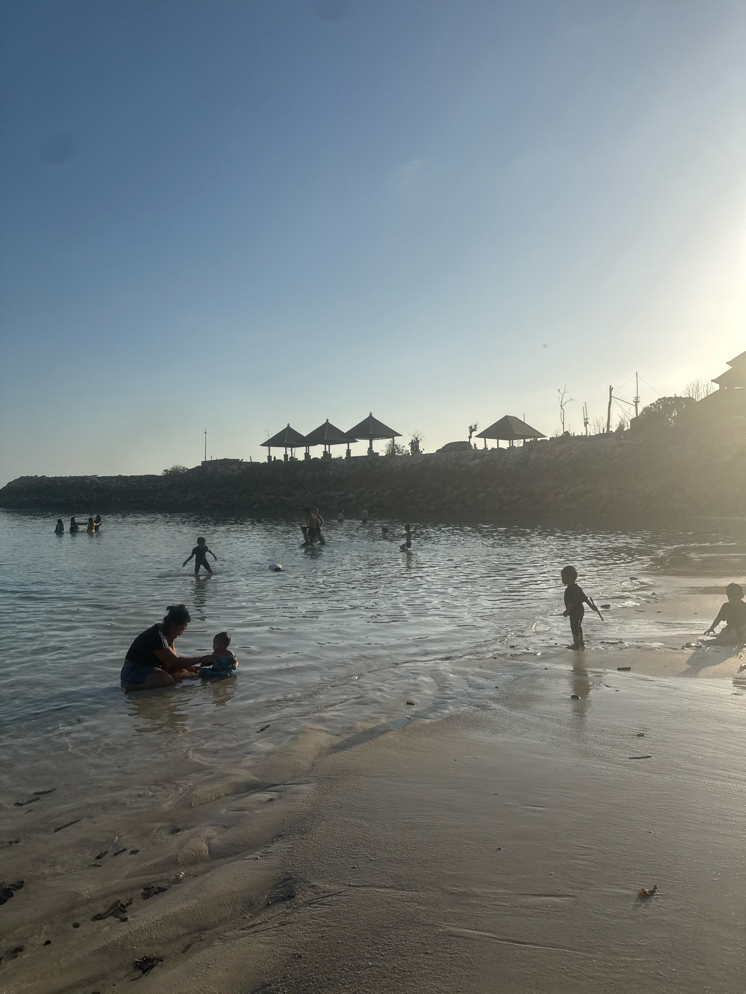 A silhouetted scene captures a beach in Kuta Selatan, Indonesia, during a sunset or sunrise. Several people, mostly children, are visible in and near the shallow water. In the foreground, a person kneels in the water with a baby. Further out, children wade and play. In the background, a line of thatched-roof gazebos is situated on a rocky embankment, with structures and trees visible behind them. The sky is clear and pale blue, with the sun creating a bright glow on the right side of the image. The wet sand near the water's edge reflects the ambient light.