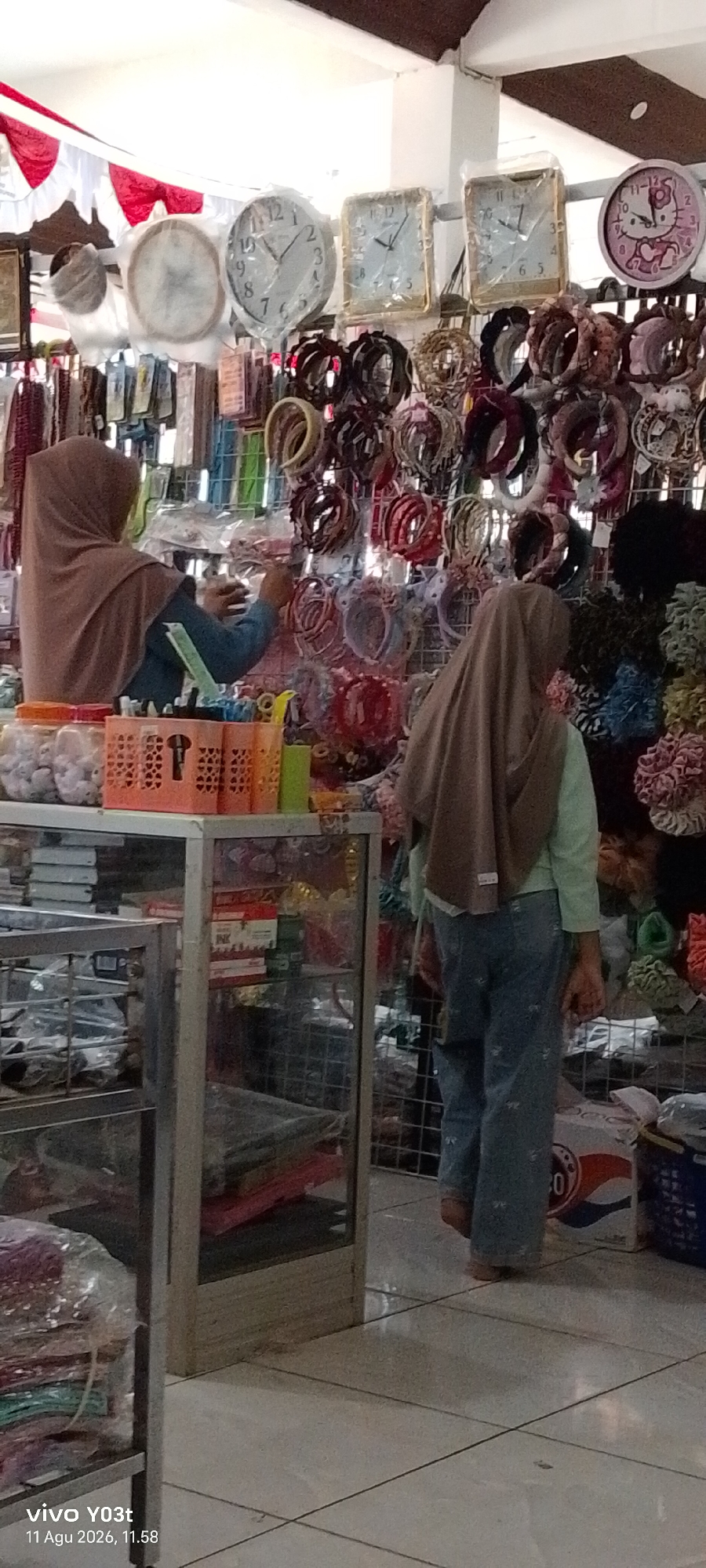 The image shows an indoor market stall, likely in Atula, Indonesia, filled with various accessories. In the foreground, two individuals are visible. One person, wearing a light brown hijab and a blue long-sleeved shirt, is attending to the stall, holding an object in their hands. The other person, also wearing a light brown hijab and a mint green shirt with blue jeans, is facing away from the camera and looking at the items on display.
The stall features a wide array of hair accessories, such as headbands and scrunchies, organized on racks. There are also several clocks displayed on the upper part of the stall, including a round analog clock and two square analog clocks, one of which is covered in plastic. In the lower part of the image, glass display cases are visible, containing what appears to be packaged goods and other merchandise. The floor is tiled with a light color. A Pepsi box is also present in the background. The overall ambiance suggests a bustling retail environment selling affordable fashion accessories.