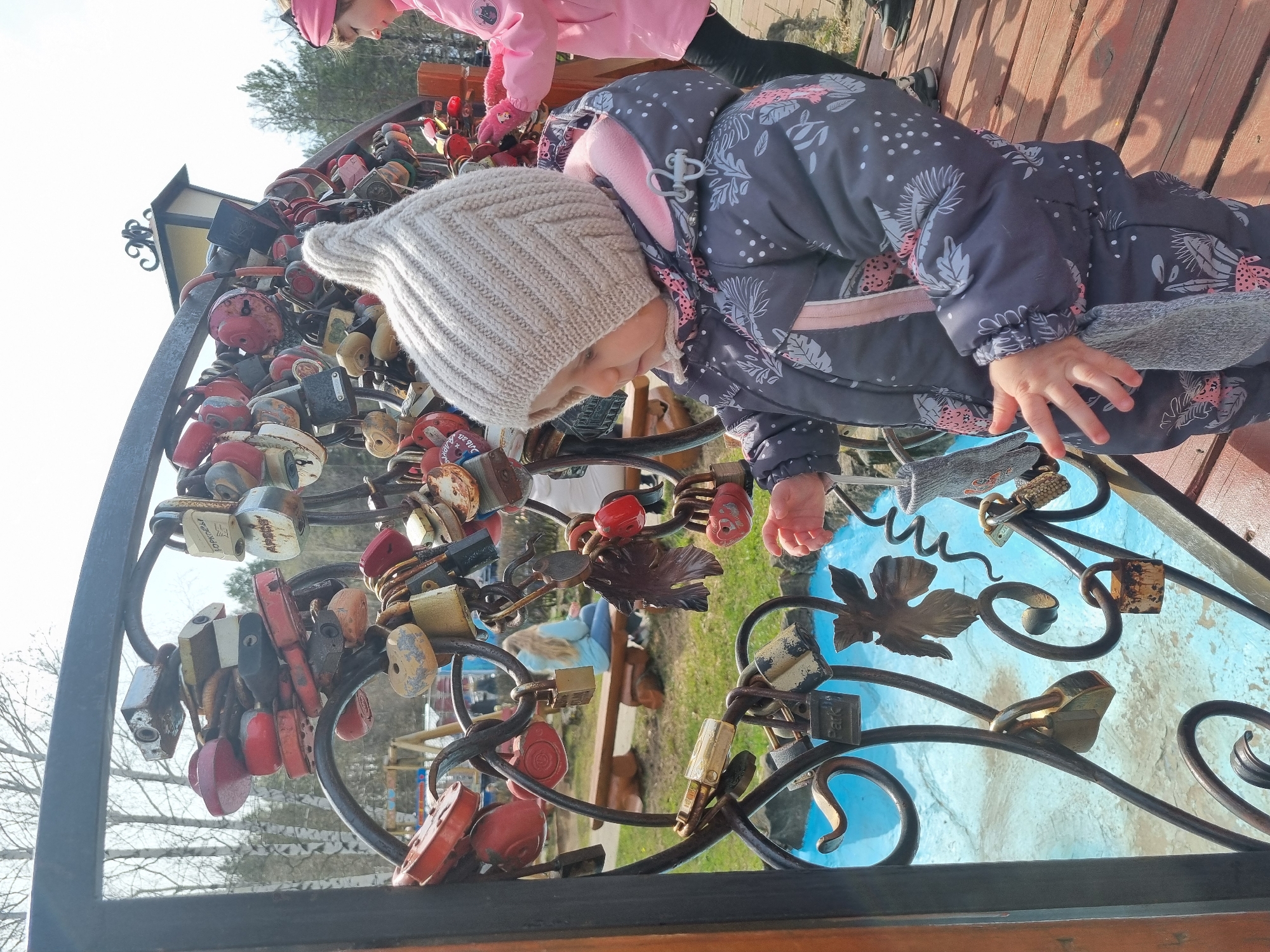 A child wearing a gray winter hat and a gray and pink patterned snowsuit is leaning over a wrought-iron fence covered in padlocks of various sizes and colors. Another child in a pink jacket is partially visible in the upper left corner. The fence is attached to a wooden structure, possibly a bridge or platform. The background includes trees and a glimpse of a bench. The scene appears to be outdoors in daylight. The location is Unknown, Unknown.
