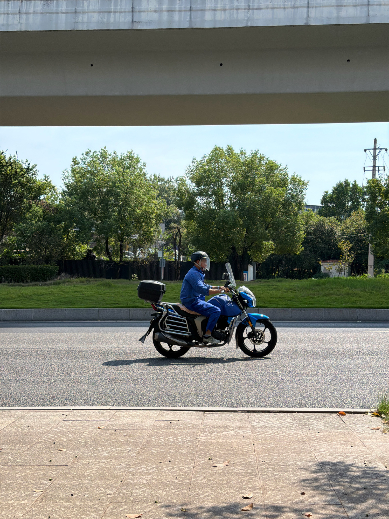 A person wearing a blue uniform and a helmet is riding a blue motorcycle with a black storage box on the back. The motorcycle is on a paved road, moving from left to right. In the background, there are trees, a grassy area, and a bridge overhead. The scene appears to be in an urban setting, possibly in Jiangjin, China, given the architectural style and vegetation. The lighting suggests it is daytime, with shadows cast on the road and sidewalk. The overall impression is one of a casual commute or delivery in a well-maintained urban environment.
