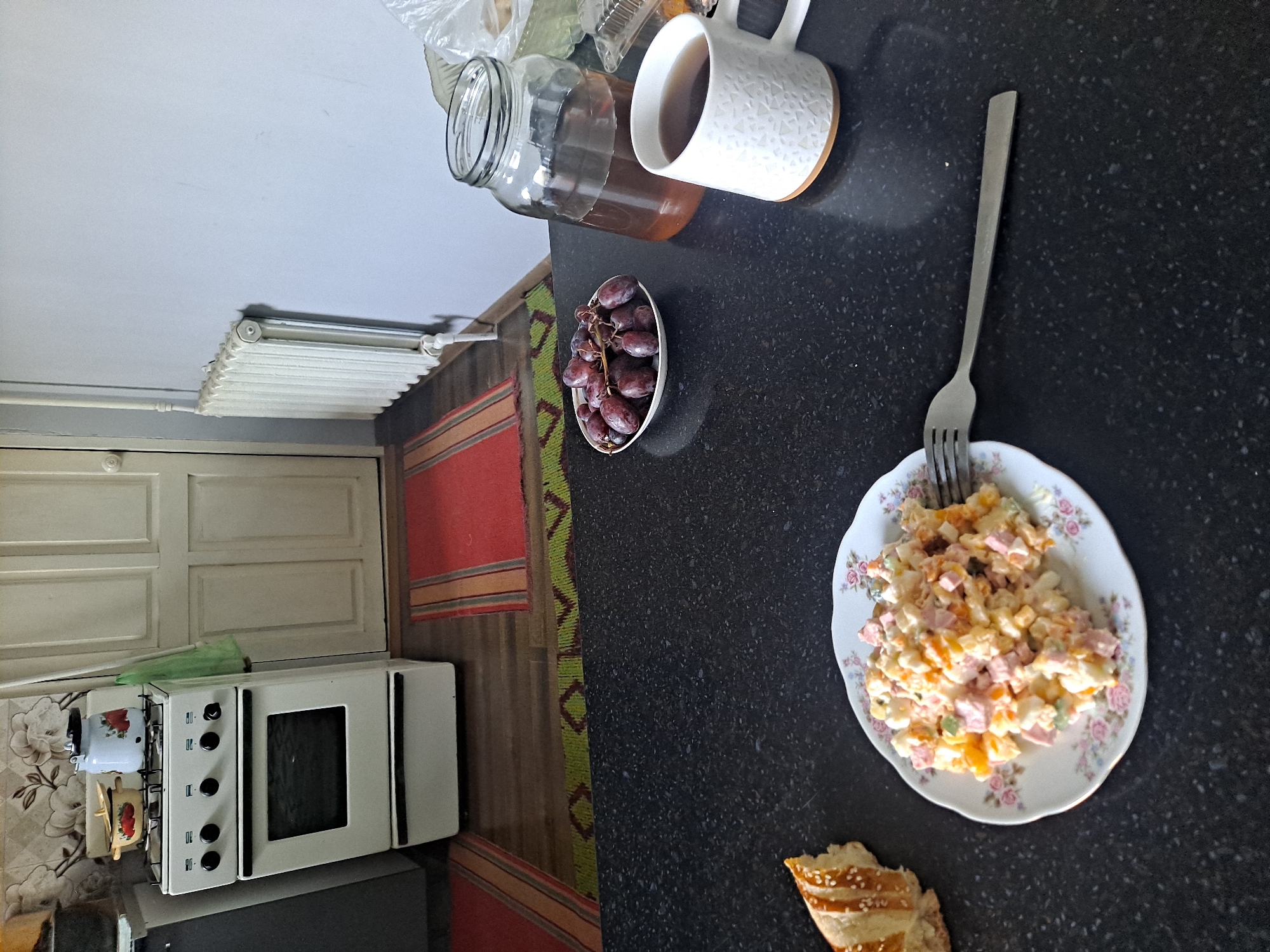 The image depicts a kitchen scene with a breakfast spread on a black countertop. In the foreground, a plate of salad and a piece of bread are visible, along with a fork. To the right, a mug of tea and a jar of liquid are placed next to a bowl of grapes. In the background, a white stove and a closed door are visible, along with a radiator on the wall. The setting appears to be a home kitchen.