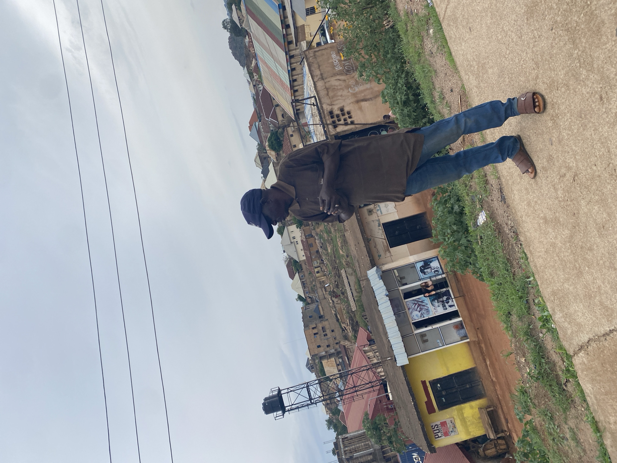 A medium-shot of a man in Gura Top, Nigeria, is positioned on a concrete walkway with vegetation growing along its edge. The man, dressed in blue jeans, a brown shirt, sandals, and a blue baseball cap, appears to be looking at or holding something in his hands. Overhead power lines extend across the left side of the frame. Behind him is a varied cityscape of buildings, some featuring corrugated iron roofs, with a water tower visible among them. The buildings range in height and color, with yellow being particularly prominent. The overcast sky suggests a cloudy day. The overall scene captures an everyday moment within the urban landscape of Gura Top.