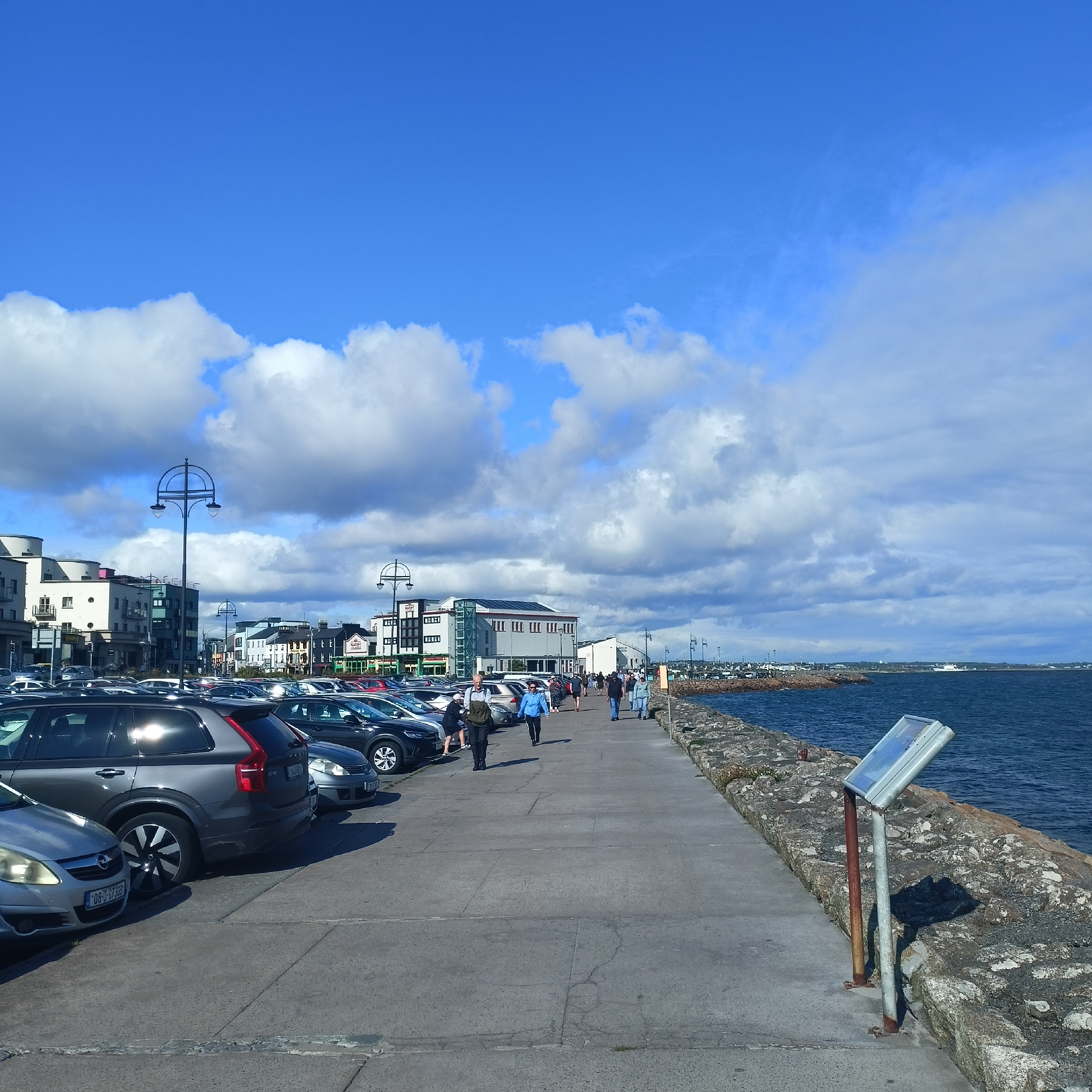 The image depicts a seaside promenade in Galway, Ireland, on a bright, partly cloudy day. In the foreground, a parking lot is filled with various cars, with a grey Volvo SUV prominently displayed on the left. Adjacent to the parking lot is a concrete promenade where a number of people are strolling. The promenade runs alongside a stone embankment that borders the deep blue, choppy waters of the ocean.
Buildings line the other side of the promenade, suggesting a commercial or residential area bordering the waterfront. These buildings feature a mix of architectural styles, including modern and traditional elements. A distinctive double-headed lamppost stands on the promenade, and other lampposts are visible further down. In the distance, more buildings and structures are visible, hinting at the extent of the town or city.
The sky is a vibrant blue, interspersed with large, fluffy white clouds that cast shadows on the ground. The sunlight is strong, creating reflections on the cars and casting distinct shadows of the people on the promenade. The presence of people walking and the parked cars suggest a typical day with activity in this coastal area. The overall mood is relaxed and pleasant, indicative of a fine day for outdoor activities by the sea. The location is identifiable as Galway, Ireland, based on the provided context. There is some text visible on the license plate of the silver Opel car: "08-D-57335".