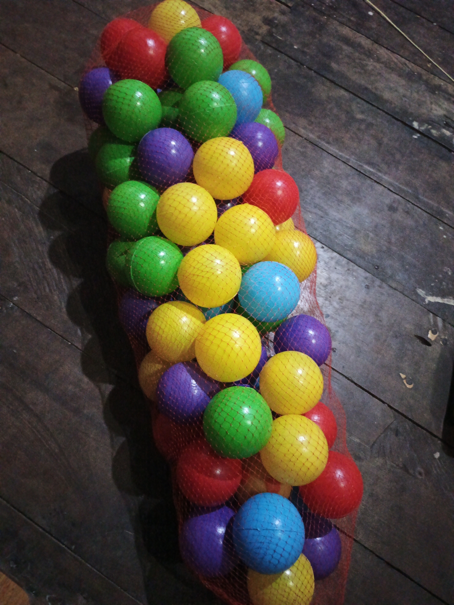 A net bag filled with colorful plastic balls, including red, green, yellow, purple, and blue, rests on a wooden floor. The bag is oriented vertically, with the balls packed tightly together. The wooden floorboards appear worn and dark, suggesting an outdoor or rustic setting. The lighting casts shadows, highlighting the texture of the wood and the spherical shape of the balls. There is no indication of people, actions, or interactions, making it a still-life composition. The scene is devoid of any specific environmental cues that would definitively place it within the city of Sinar Bulan, Indonesia, or any other specific geographic location.