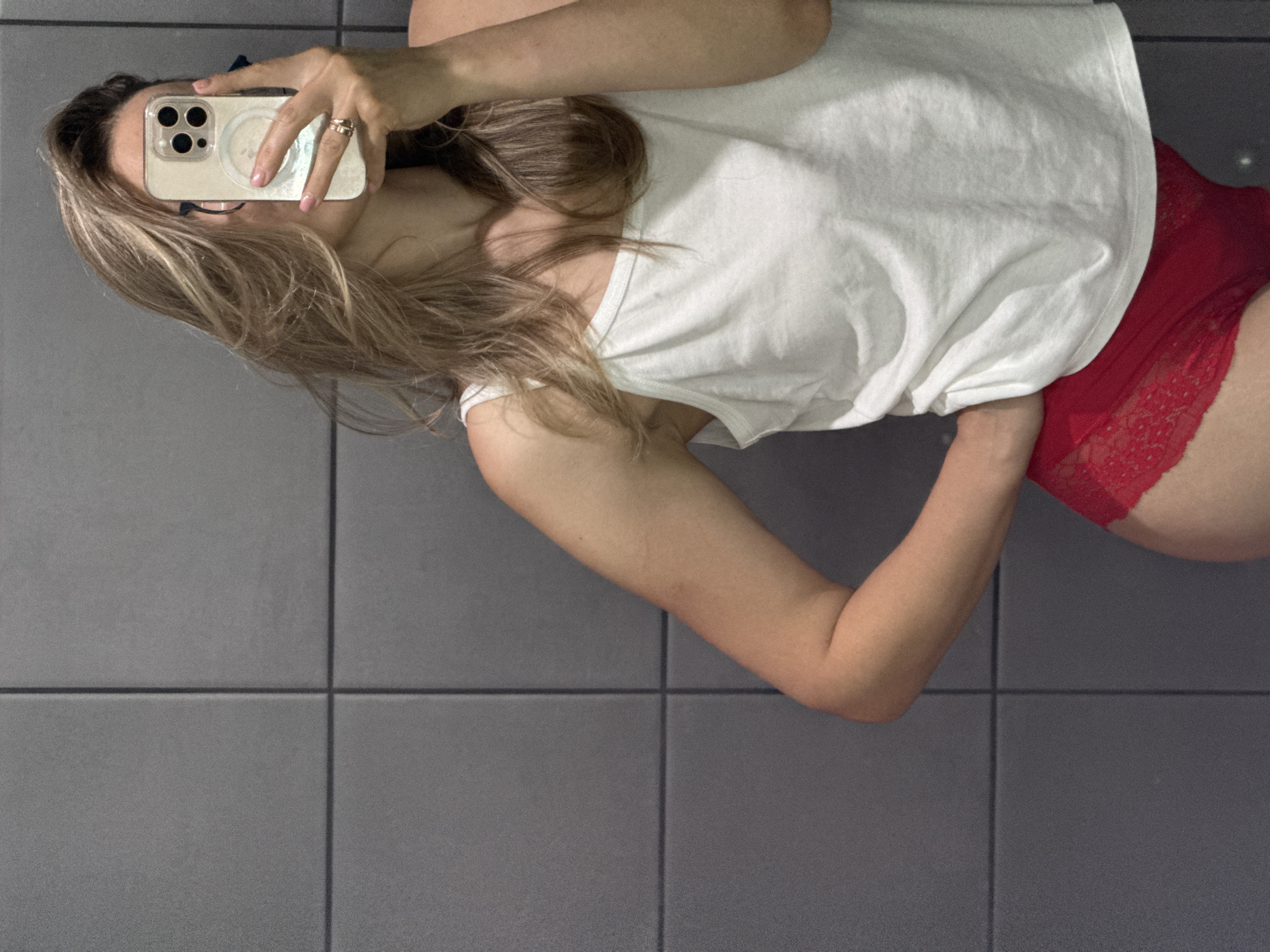 A woman with blonde hair and fair skin is taking a selfie in a bathroom. She is wearing a white tank top and red lace underwear. The bathroom walls are tiled with grey squares. She is holding a smartphone in her right hand and her left hand is on her hip. The image appears to have been taken indoors, likely in a residential bathroom. There is no indication of the city of Chaikovsky, Russia, or any other specific location.