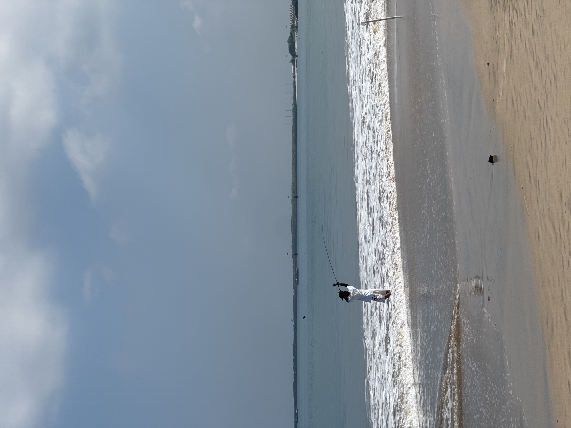 The image, originally rotated, depicts a bright daytime scene on a sandy beach. A lone person, dressed in light-colored clothing (possibly white) with dark accents on their upper body, stands in the shallow breaking waves of the ocean. They are actively engaged in fishing, holding a long fishing rod that extends out over the water.
The ocean stretches across the middle ground of the image, displaying light blue-green hues that lighten towards the horizon. Gentle white, foamy waves lap at the sandy shore where the person is standing. The beach itself is a wide expanse of light brown and tan sand, showing ripples and wet patches from the receding tide. In the distance, a low-lying coastline is visible, appearing as a thin strip of land with faint indications of structures or vegetation under a clear, bright sky. The sky above is a pale blue, with soft, scattered white clouds, suggesting fair weather.
The scene conveys a peaceful outdoor activity, likely in the morning or afternoon given the clear light. The location is consistent with a coastal area like Kuta Selatan, Indonesia. There is no visible text in the image.