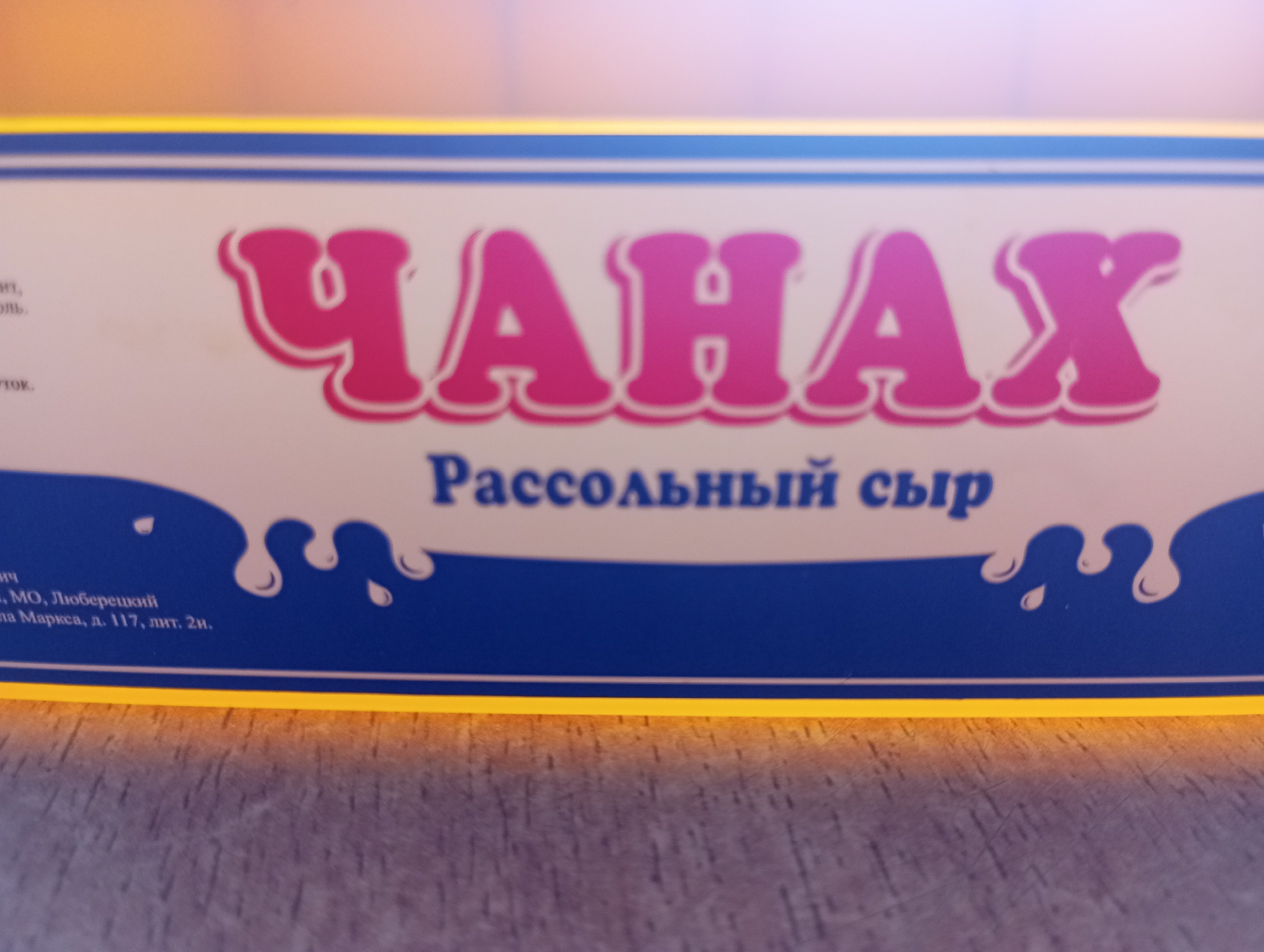 The image shows a close-up of a blue and white box with yellow trim. The box has pink lettering that says "ЧАНАХ" which translates to "CHANAH" in English. Below this, in smaller blue text, it says "Рассольный сыр," meaning "brined cheese." The text and design are on the front of the box. The background is blurry, showing a wooden surface. There is no information in the image that provides any context about the city of Kirzhach, Russia.