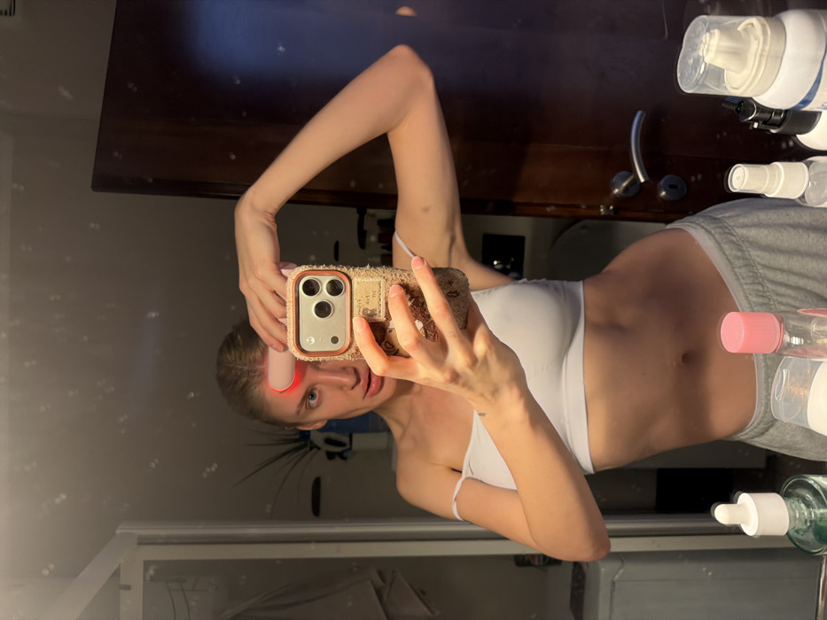 The scene depicts a female individual taking a mirror selfie in a bathroom, likely within a private residence or hotel in Budva, Montenegro. The woman, with light hair pulled back, is reflected in the mirror, wearing a white strapless or off-the-shoulder crop top and grey sweatpants. She holds a light-colored smartphone with a textured, possibly tweed or sequined, case in her right hand, positioned to capture her reflection. Her left hand holds a pink and white beauty device with a glowing red light emitter against her forehead. Her mouth is slightly open, and her gaze is directed towards the phone camera.
The bathroom environment reflected in the mirror shows a dark wooden door with a silver handle and lock mechanism on the right side. To the left, a portion of what appears to be a washing machine or large laundry basket is visible. A dark plant in a light-colored pot is partially visible in the upper left background. The foreground, along the bottom edge of the mirror, displays a collection of personal care products on a counter, including bottles with white, pink, and clear liquid, pump dispensers, and spray bottles. The overall lighting is dim, with direct light sources reflecting as bright spots in the mirror, particularly around the woman's head and the beauty device.