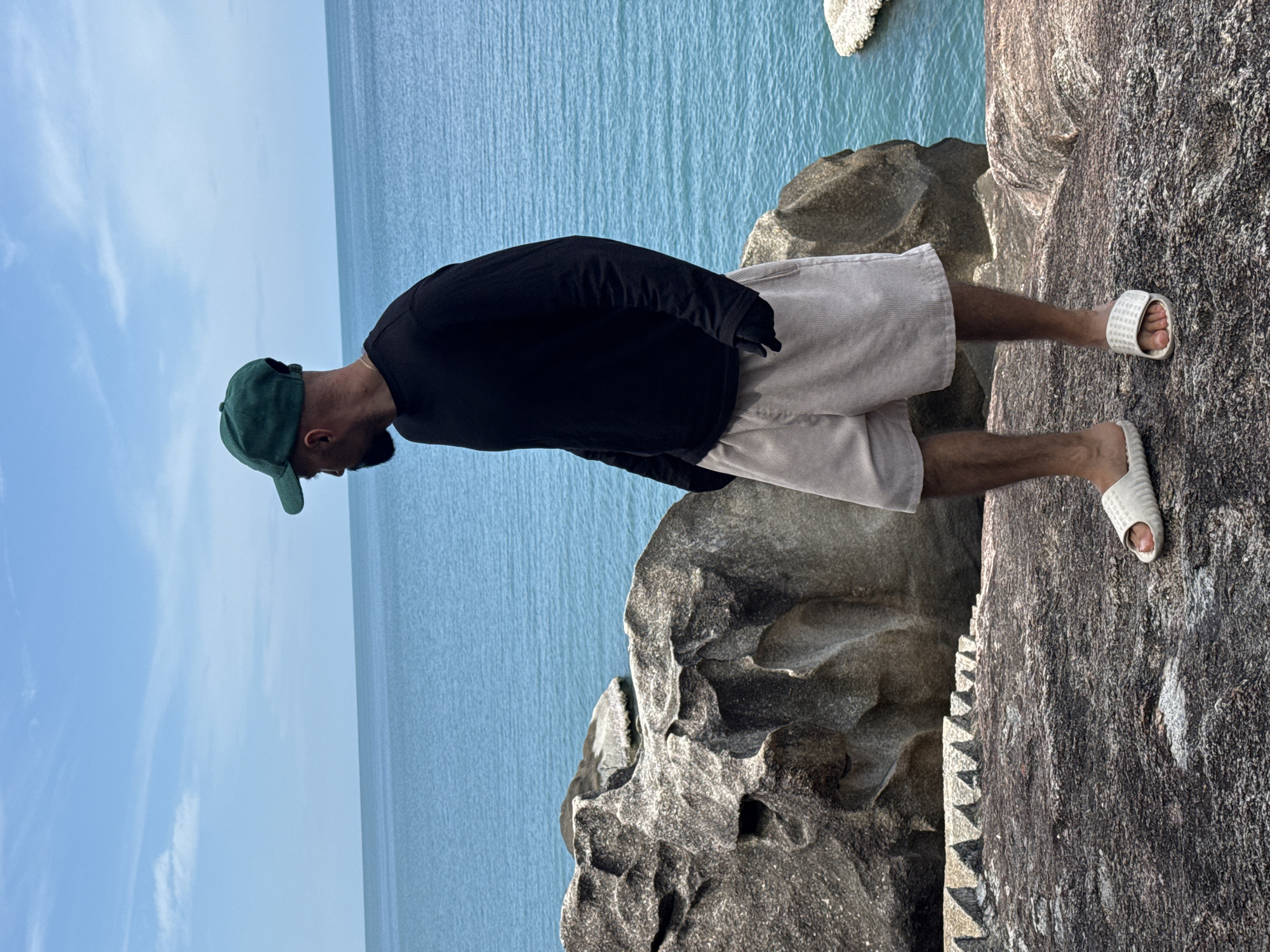 A person wearing a black long-sleeved shirt, light beige corduroy shorts, a green baseball cap, and white slides stands on a rocky surface overlooking the ocean. The person is facing to the right, looking out at the blue water. In the background, large rocks are visible along the shoreline, and the sky is a clear blue with a few wispy clouds. The location is the coast of Ko Samui District, Thailand, known for its granite rock formations.