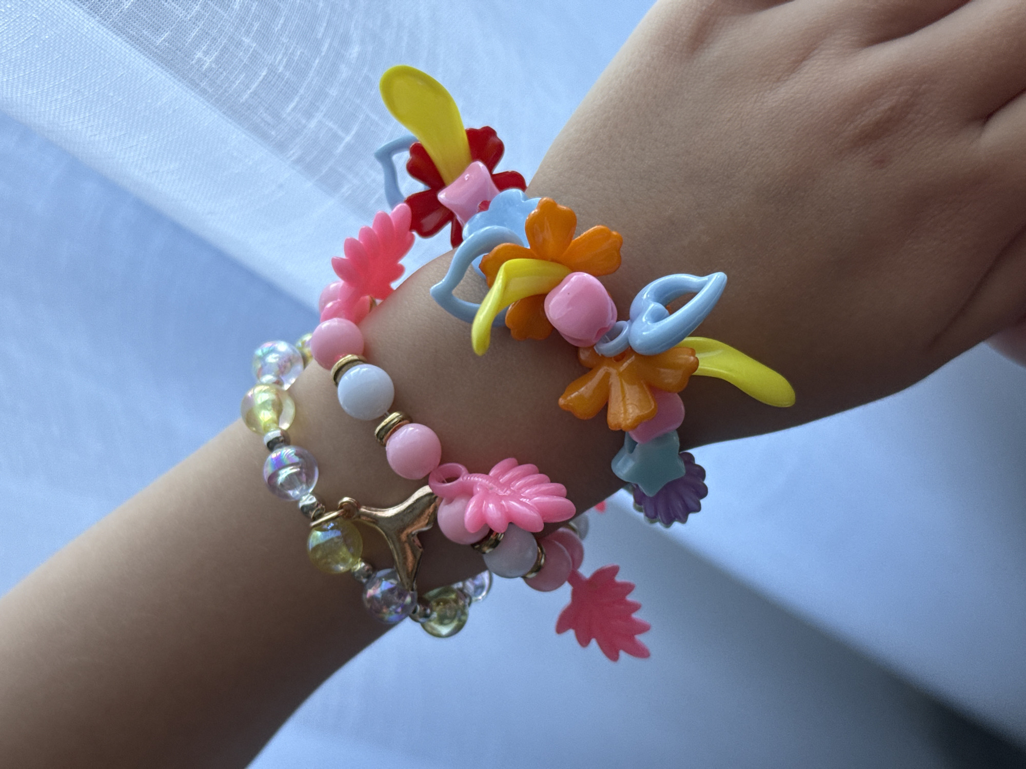This close-up image focuses on a child's arm and hand, adorned with multiple colorful bracelets, set against a softly lit background. The lighting is bright and natural, indicating daytime.
The arm, with a smooth skin tone typical of a young person, serves as the central subject. Several bracelets are worn on the wrist. One prominent bracelet features an assortment of brightly colored plastic shapes, including flowers and abstract designs, in vivid yellow, red, light blue, orange, and pink. A smaller, pale pink leaf-shaped plastic charm is also part of this group or dangles near it. Another bracelet, worn closer to the hand, consists of round beads in white, soft pink, and clear iridescent spheres that show subtle rainbow reflections. This beaded bracelet also incorporates small golden spacer beads and a unique golden, abstract charm that resembles a stylized horn or crescent.
The background is a soft, out-of-focus expanse of light blue and white, displaying faint textures consistent with a sheer or translucent fabric, likely a curtain, suggesting an indoor setting near a window. The primary activity depicted is the wearing of these decorative items. No text is visible in the image. The overall impression is one of childlike adornment and cheerfulness due to the vibrant color palette. The location context provided is Murom, Russia.
