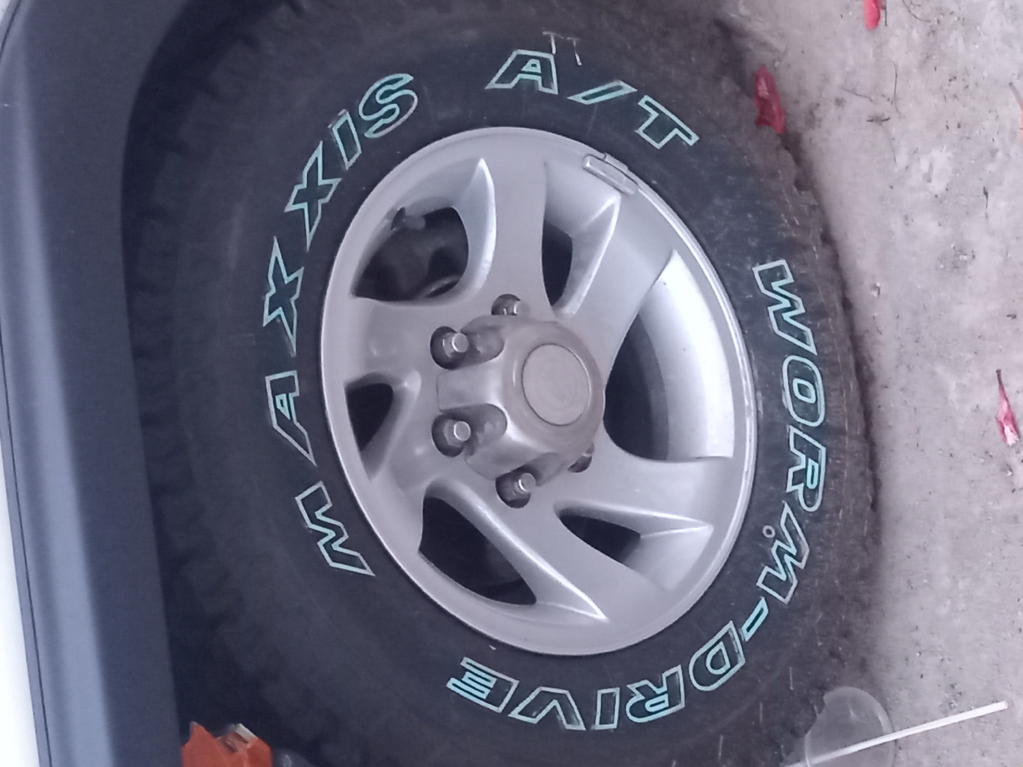 A close-up, slightly angled shot shows the side of a car tire. The tire is black with light blue lettering. The words "MAXXIS" are visible on the top half of the tire, and "A/T DRIVE" and "WORM-D" are visible on the bottom half. The tire is mounted on a silver alloy wheel with five spokes. A portion of the car's dark gray fender flares is visible at the top of the image. The ground appears to be a mix of dirt and small gravel. The overall lighting is natural and the scene is static. There are no people visible in the image. The image context does not provide any specific location within Đà Nẵng, Vietnam.