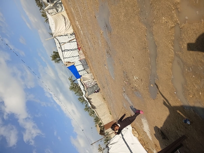 Tents stand in a muddy landscape under a partly cloudy sky. A child in dark clothing and pink shoes lies near a white tent. Patches of standing water cover the ground. A shadow stretches across the mud, suggesting the presence of a person taking the photograph. Trees are visible in the background behind the tents.