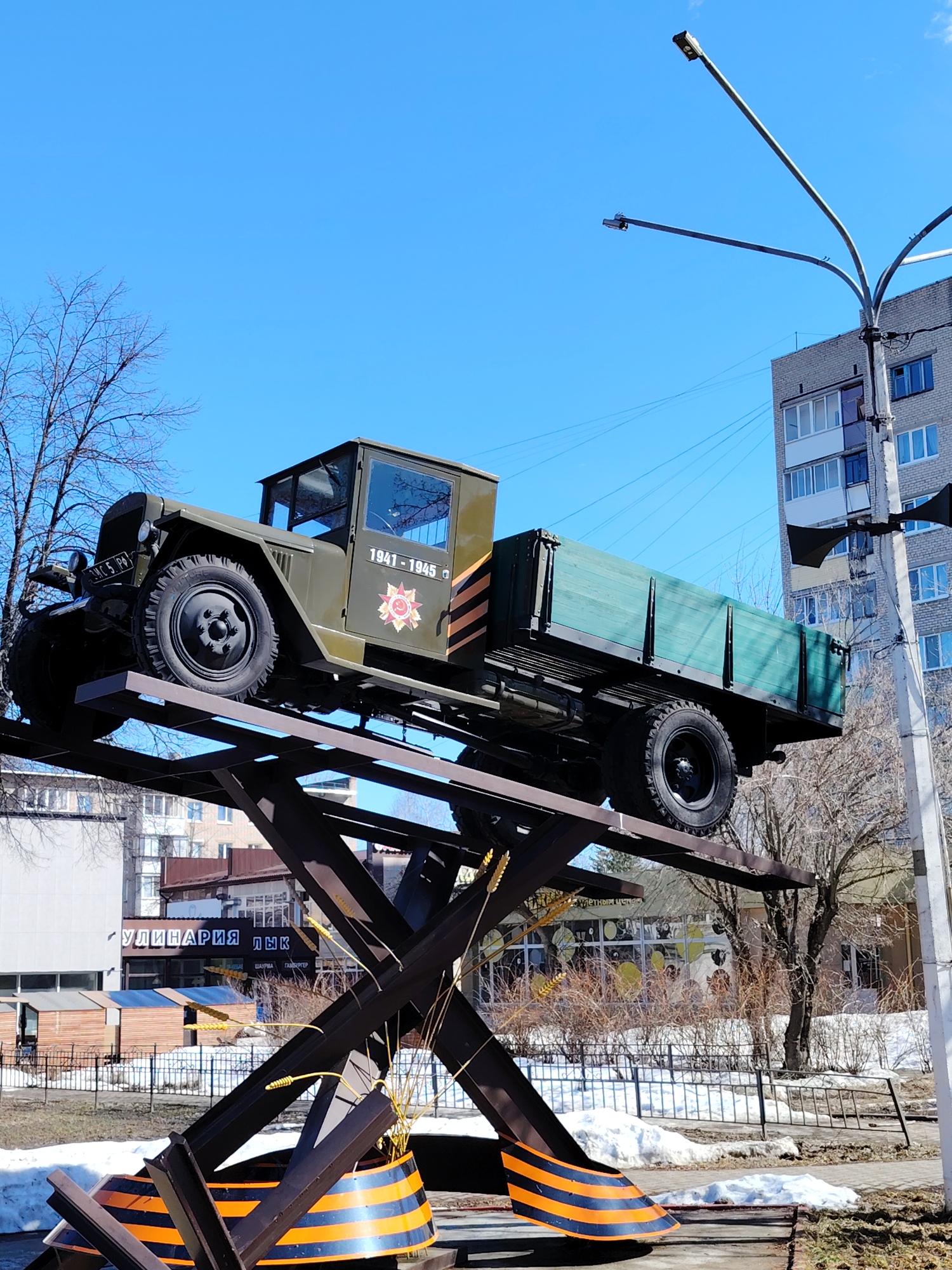 The image shows a ZIS-5 truck monument in the city of Chaikovsky, Russia. The truck is painted olive green and rests on a metal structure decorated with the St. George ribbon in black and orange stripes. The cab of the truck has the text "1941-1945" and a red star emblem. The monument is located in a public space with buildings and trees in the background. Snow patches remain on the ground, indicating a late winter or early spring setting. A street light pole stands adjacent to the monument. A building with the signage "УЛИЛНАРИЯ ЛЫК" can be seen in the background.