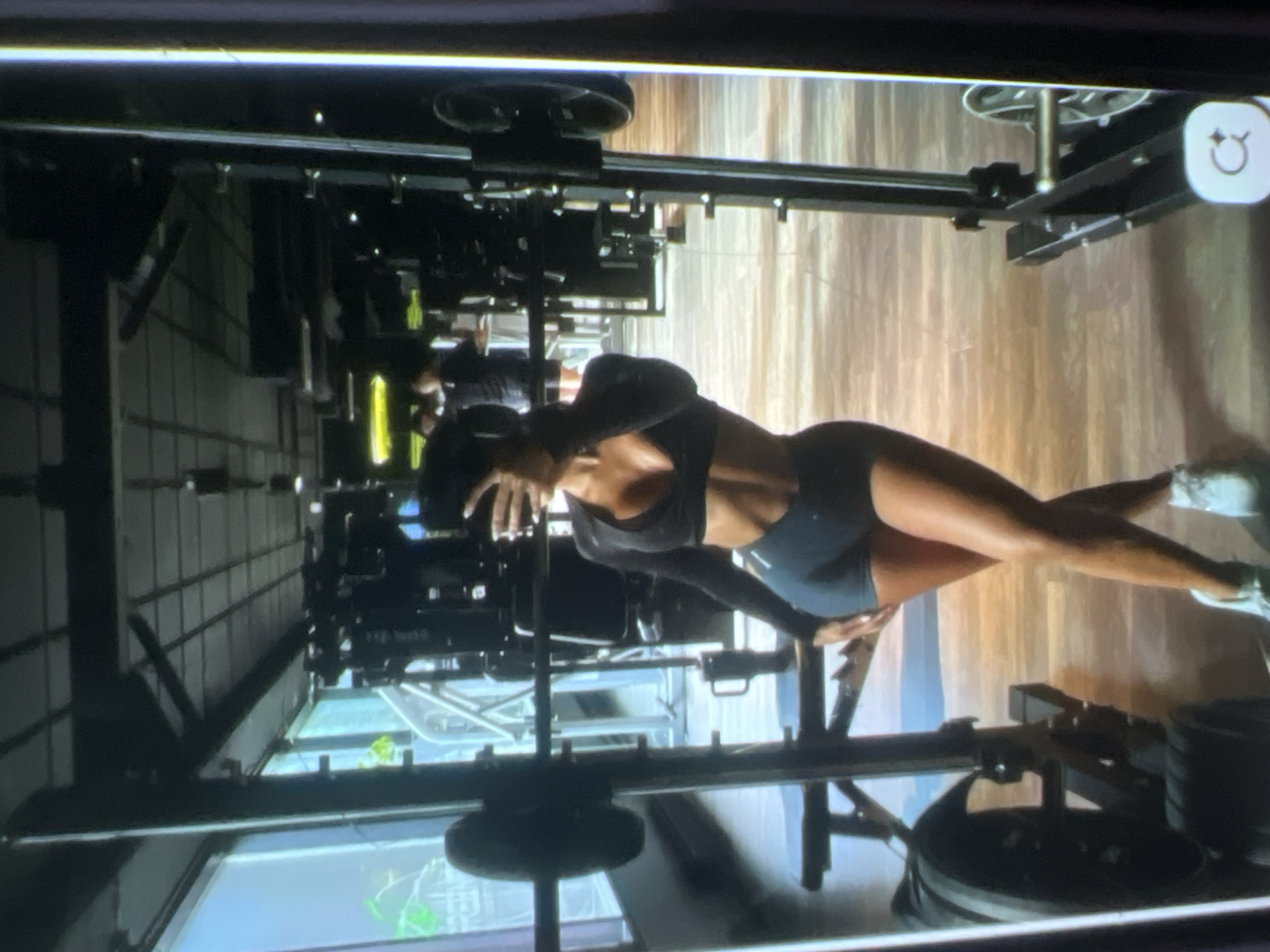 An individual, likely female based on attire, is captured in a full-length mirror selfie within a gym. They are wearing a black long-sleeved crop top and dark athletic shorts, accentuating their abdominal muscles. The individual's legs are crossed, and they are holding a smartphone to take the picture. The gym environment is visible in the background, featuring weightlifting equipment, including a barbell rack with weights on either side. The flooring appears to be wood-effect laminate. The lighting suggests an indoor setting, possibly during daylight hours due to reflections from large windows on the left side of the image. The overall scene conveys a focus on fitness and personal training. The location is specified as the city of Kara-Dıykan, Kyrgyzstan.