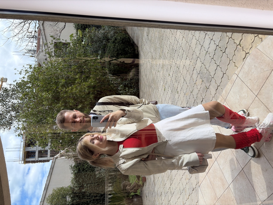 An image captured as a reflection in a glass surface depicts two individuals, a female and a male, embracing and smiling. The female, positioned closer to the reflective surface, is holding a light-colored smartphone in her right hand, indicating she is taking the photograph. She has blonde hair styled in pigtails and is dressed in a light-colored bomber jacket over a red top, a white skirt, light hosiery, and white sneakers with pink accents, complemented by mismatched leg warmers (pink on the right leg, red on the left). A small, light-colored handbag hangs from her left shoulder. The male stands behind her, wearing a light-colored hooded jacket, light blue jeans, and dark sneakers, with a dark backpack on his shoulders. Both individuals have their arms around each other's waists.
The scene reflected beyond the individuals shows an outdoor area. The ground consists of light-colored, patterned pavers, likely concrete, leading away from the reflective surface. Lush green foliage, including bushes and trees, lines the paved area. In the background, a multi-story building under construction, characterized by exposed concrete framework and rebar, is visible under a partially cloudy blue sky. A yellow crane boom extends above this construction site. To the right of the construction, a white building with a sloped roof is partially obscured by trees. The architecture and ongoing construction are consistent with residential or mixed-use development within the city of Budva, Montenegro, though a precise address is not discernible from the visual evidence. The immediate foreground shows light brown tiles on the floor where the reflection is occurring.