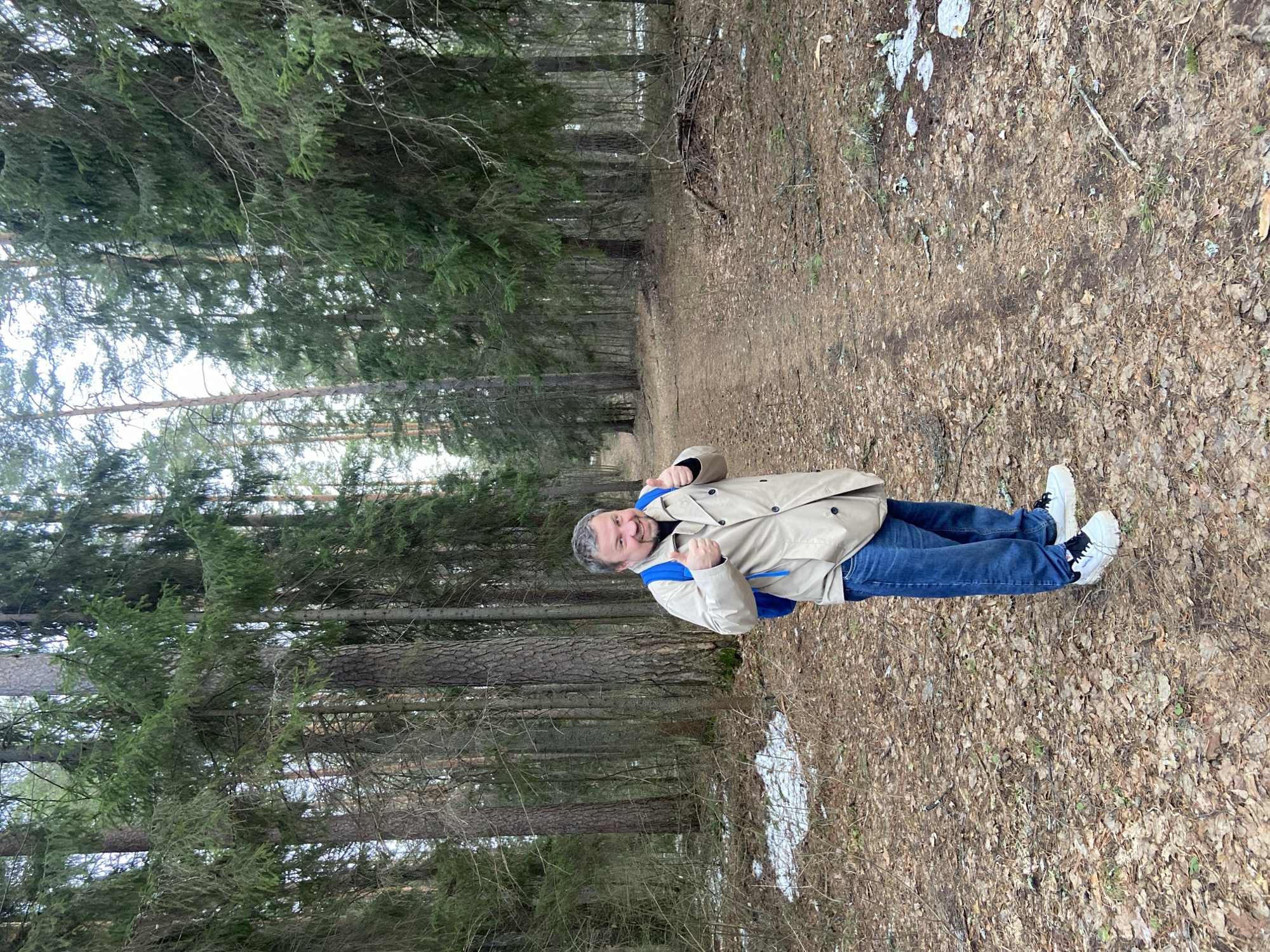 A man stands on a leaf-covered path within a dense forest. He wears a tan trench coat, blue jeans, and white sneakers, and has a blue backpack. The forest consists of tall evergreen trees, potentially spruces, and some bare deciduous trees. Patches of snow are visible on the ground near the edge of the path. The location might be near Noviy, Russia, but this cannot be definitively confirmed from the image alone. He is smiling and giving two thumbs up.