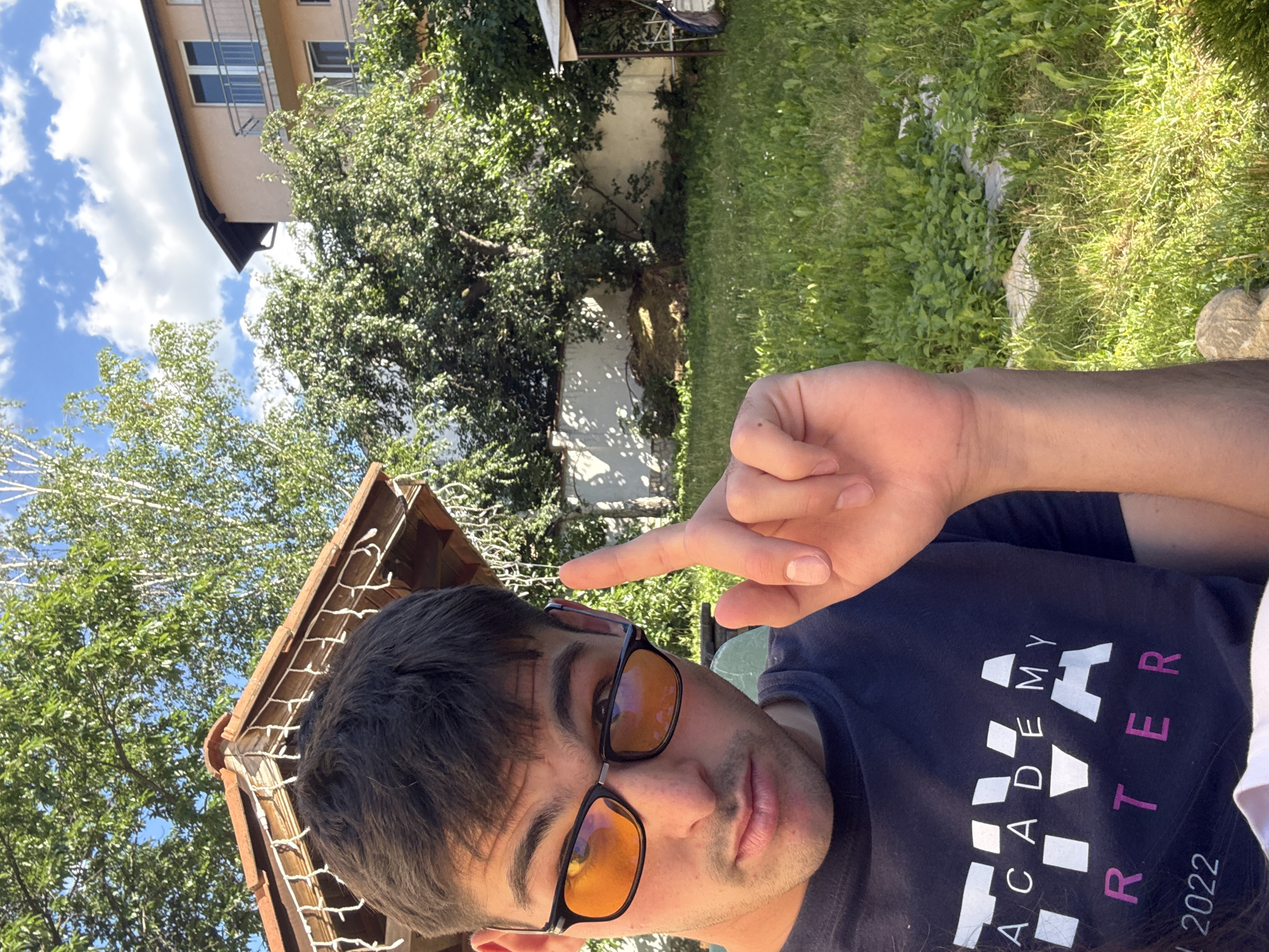 A young man with dark hair and glasses is taking a selfie outdoors. He is wearing a dark t-shirt with white and pink lettering. His right index finger is raised. The background shows a yard with green grass, trees, and a building. The man's t-shirt has the words "ACADEMY" and "ARTER 2022" printed on it. He is looking at the camera with a neutral expression. The scene appears to be in Sofia, Bulgaria, given the architectural style of the building and the general environment.