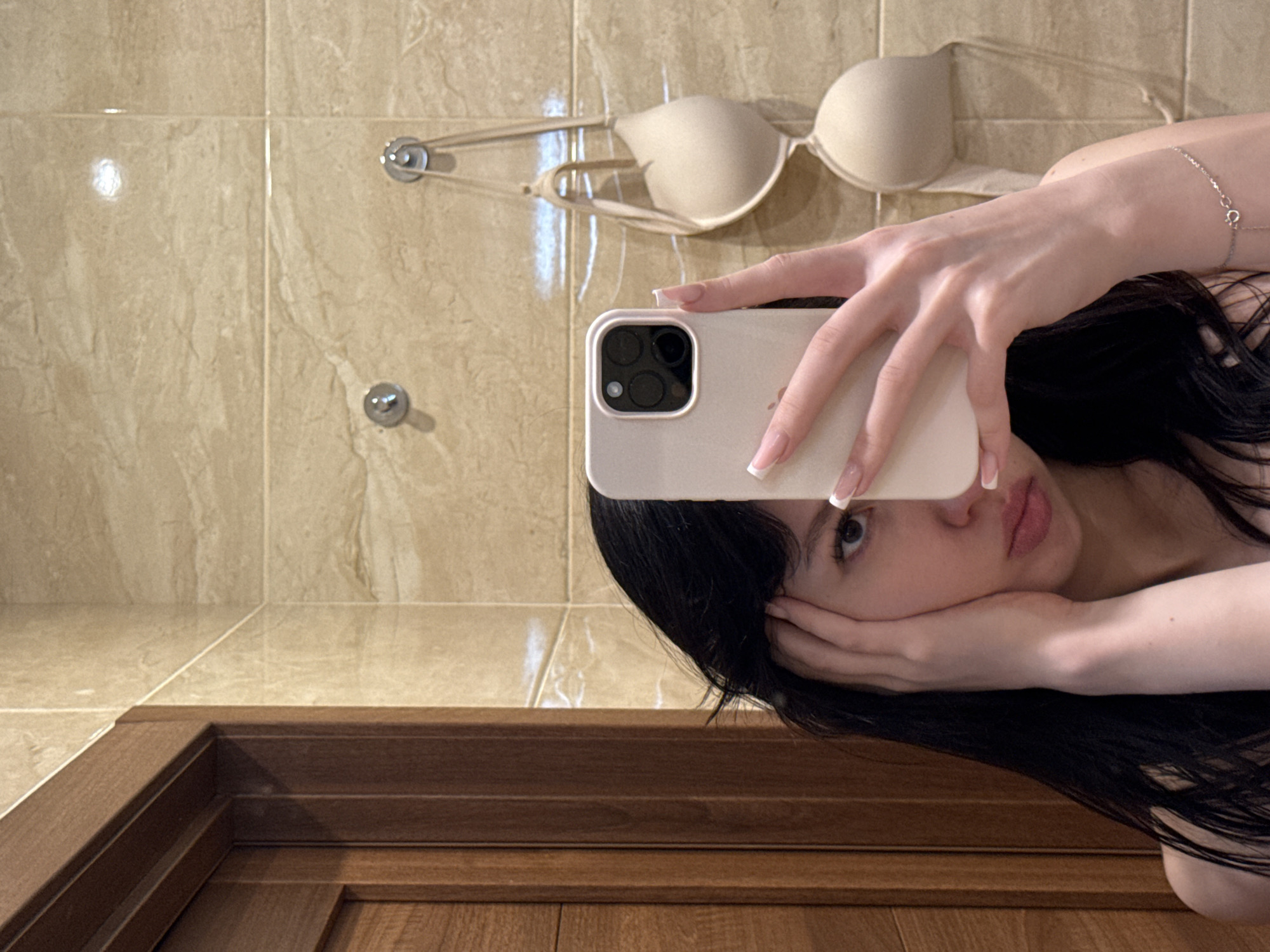 The image is a selfie taken in a bathroom. The subject is a young woman with dark hair and fair skin, holding a smartphone in her right hand to take the picture. She is looking at the camera with a neutral expression, with one hand resting on her face. A beige bra is hanging on hooks on the tiled wall behind her. The bathroom features marble-patterned tiles and a wooden door frame. There is a silver bracelet on the woman's left wrist. The image does not provide any information about the city of Khimki, Russia.