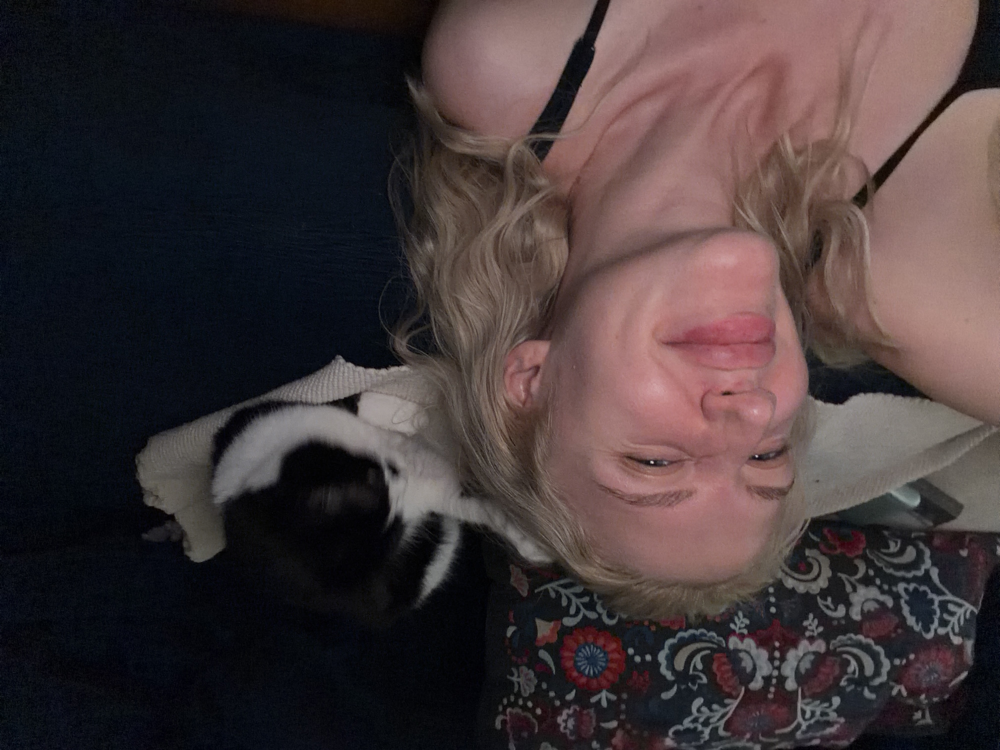 A woman with blonde, wavy hair is lying on her back, likely on a couch or bed, with her head tilted and smiling. A black and white cat is resting on her head or near her, partially obscured. The background appears to be a dark, possibly indoor setting. There is no information in the image to suggest a location in Belgrade, Serbia.