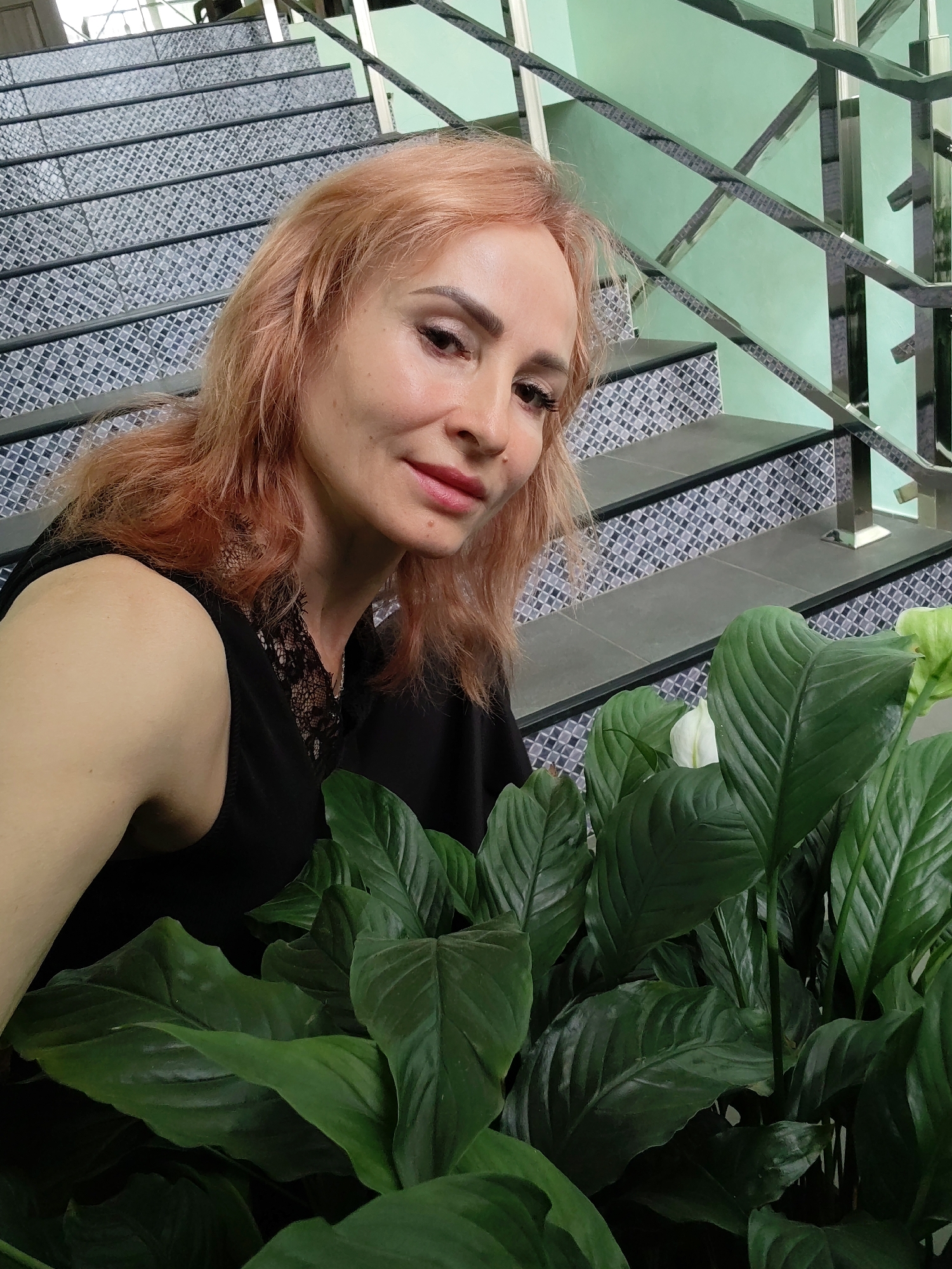 A Caucasian woman with fair skin and reddish-blonde hair is positioned in the foreground, partially obscured by large green leaves of a peace lily plant. She is looking towards the camera with a slight smile, her makeup including lipstick and eye makeup. She is wearing a black sleeveless top with lace detailing at the neckline. The background shows a staircase with patterned tiles and a metal railing against a light green wall. The scene appears to be indoors, possibly in a public space like a building lobby or a staircase area. There are no recognizable landmarks or indications that place this image specifically in Yoshkar-Ola, Russia.