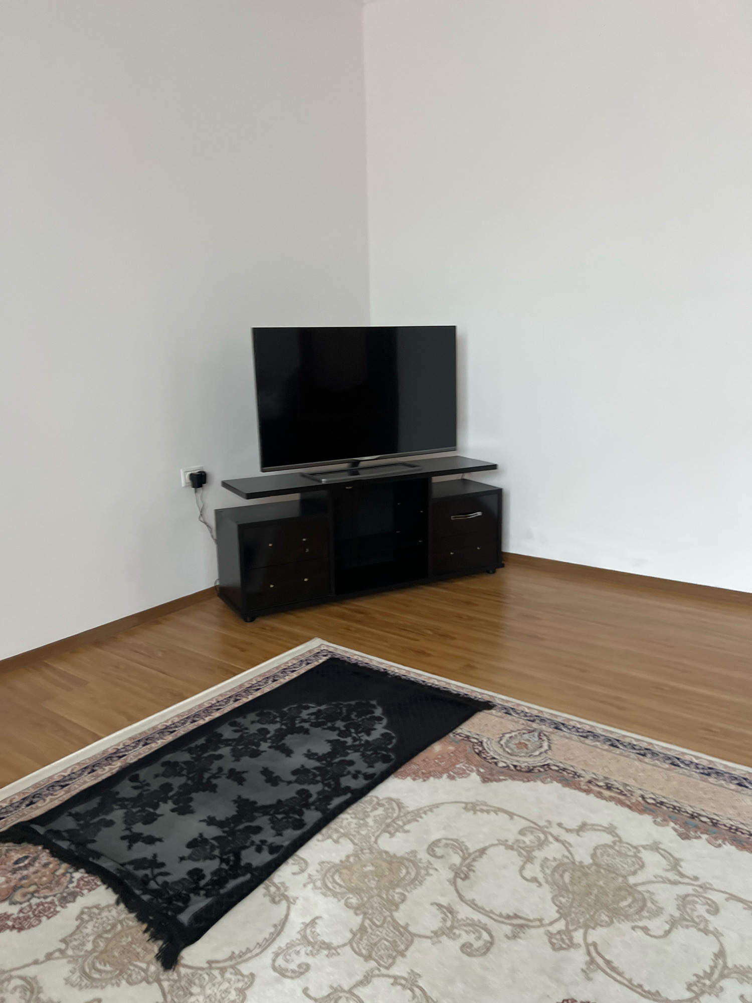 The image shows a corner of a room with white walls and a wooden floor. A black television is mounted on a black TV stand with drawers. A black prayer rug with a floral pattern is placed on top of a larger, beige rug with an ornate design. A power outlet and plug are visible on the wall behind the TV stand. The scene appears to be a domestic setting in Novogagatli, Russia.