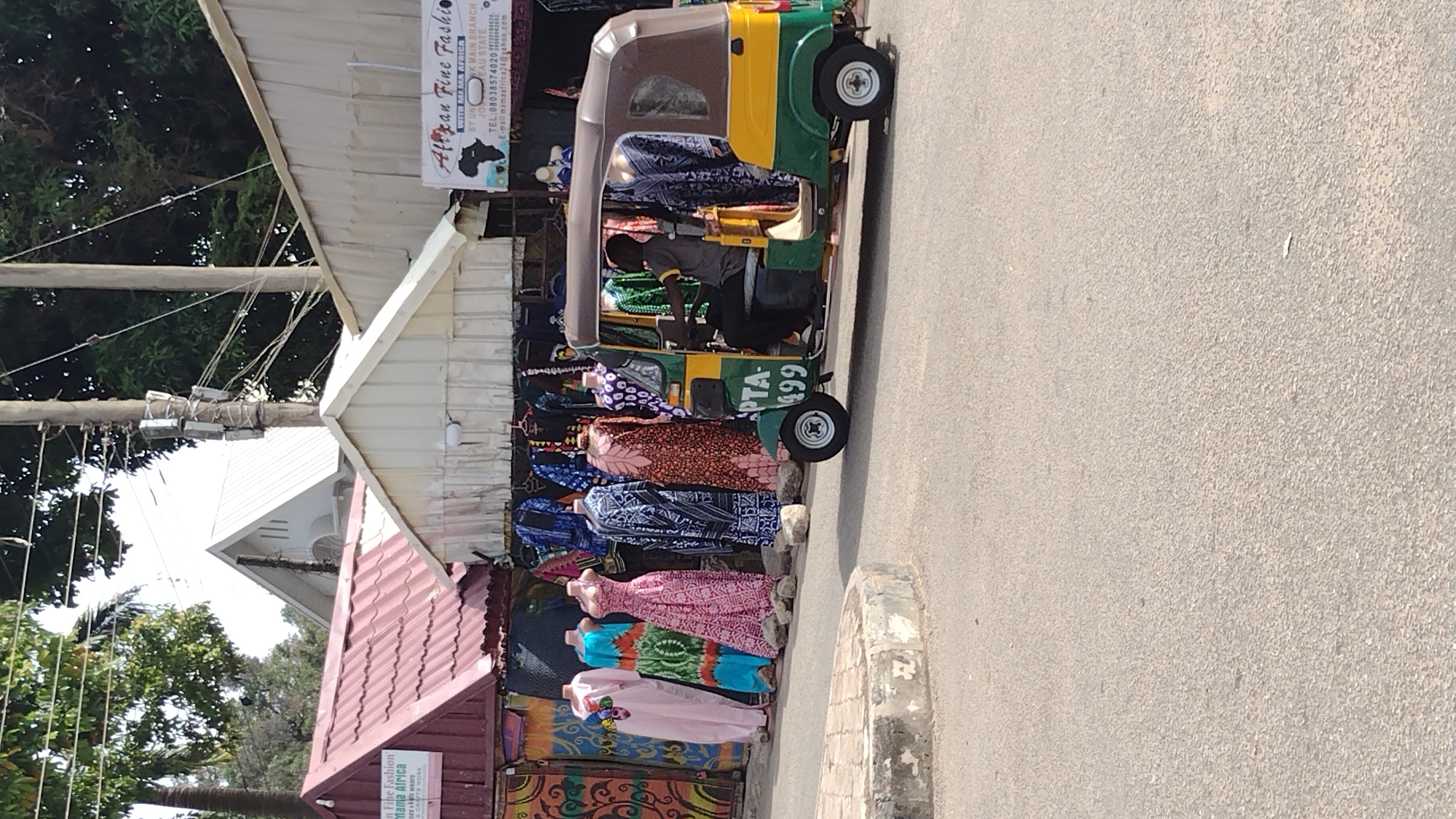 Here is a description of the image:
The image shows a roadside shop in Jos, Nigeria called "African Fine Fashion." The shop sells Mama Africa textiles and features mannequins displaying colorful dresses and fabrics. A "keke," or auto-rickshaw, is parked next to the shop. A driver is visible inside the vehicle. The street is paved. Overhead power lines are evident. The sky is bright.