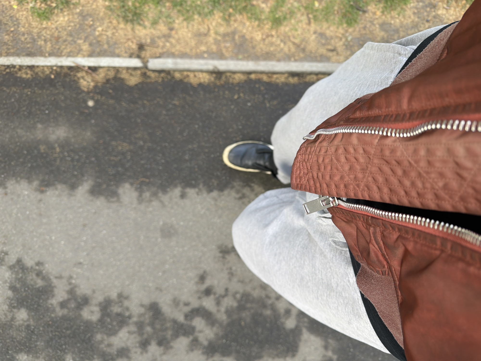 The image captures a top-down, first-person perspective of a person standing or walking outdoors. The primary subjects are the individual's clothing and footwear, seen from the waist down. The person is wearing a rust-colored, possibly quilted or textured jacket, featuring prominent silver zippers. Below the jacket, light grey sweatpants are visible, and on the left foot, a dark shoe with a cream-colored sole.
The scene depicts an outdoor setting, likely a sidewalk or paved path next to a grassy area. The ground is dark asphalt or pavement, showing some lighter, irregularly shaped patches that could indicate dampness, wear, or dried water marks. A light grey concrete curb runs horizontally across the upper part of the image, separating the pavement from a strip of dry, light brown grass or dirt. Beyond this strip, green foliage, possibly a lawn or bushes, is visible, albeit out of focus.
The lighting suggests it is daytime, but no specific time of day can be determined. There is no visible text in the image. The overall impression is casual, capturing an everyday moment in an urban or suburban environment, consistent with the provided location context of London, United Kingdom.