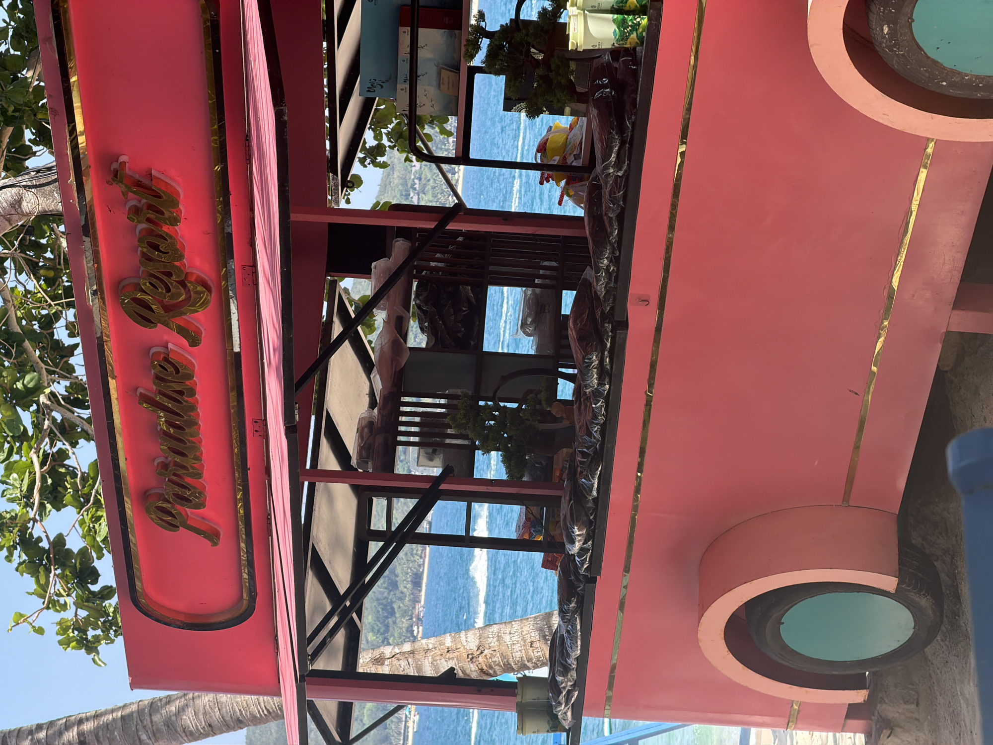 The image shows a pink structure with the words "Paradise Resort" on the side, under what looks like a canopy or awning, at an angle, with a view of the ocean and greenery visible through the structure. The scene appears to be at Nam Nha Trang Ward, Vietnam, based on the vegetation, architecture, and coastal setting. The structure appears to be a modified double-decker bus converted into a bar or small restaurant. There is vegetation partially obscuring the sign, suggesting it's located near trees or foliage. The viewpoint is from below, looking upwards toward the structure and the sea.