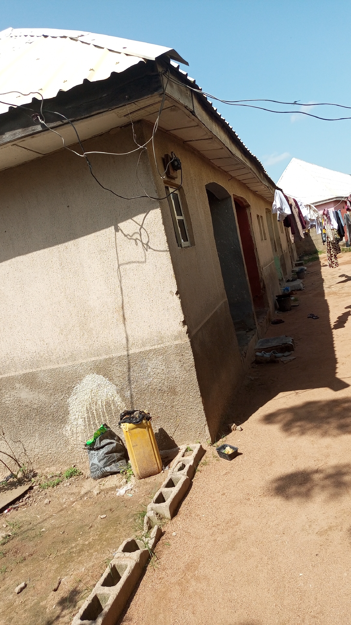 The image depicts an outdoor scene of a row of modest buildings under a clear blue sky, suggesting daytime. The buildings have corrugated metal roofs and beige, textured walls, with visible electrical wiring running along the eaves. In front of the buildings, a dirt path is present, with various objects scattered around.
To the left, a yellow plastic container and a black trash bag filled with debris are visible near the building's wall. A short line of concrete blocks runs along the dirt path. Further down the row of buildings, clothes are hung out to dry on a line, indicating a residential setting, likely in a community where this is common. Some buckets and shoes are also placed outside the doorways of the buildings. A small black tray containing what appears to be food is situated on the ground near the concrete blocks.
The overall impression is that of a simple, lived-in environment. The bright sunlight and shadows indicate a sunny day, possibly in the afternoon, given the length of the shadows. There is no discernible text in the image. The scene appears to be set in Jalingo, Nigeria, as per the provided location context, suggesting a warm, possibly tropical climate. There are no people clearly visible in the frame, but the presence of clothes drying and personal items implies human habitation.