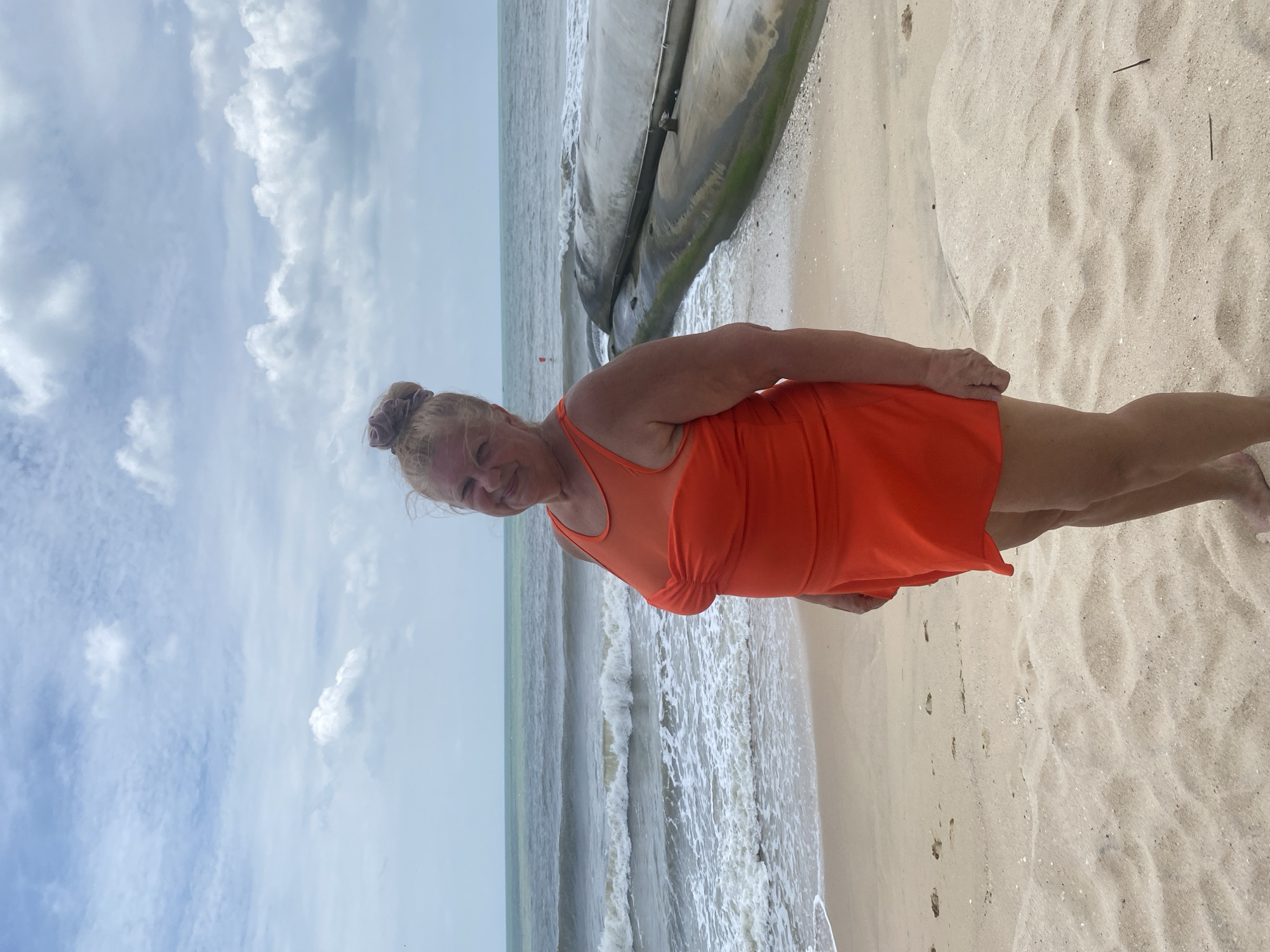 A woman is standing on a sandy beach, facing the camera and smiling. She is wearing a bright orange swimsuit. The ocean with waves is behind her, and the sky is cloudy. To her right, there are large, grey, cylindrical objects partially submerged in the sand near the water's edge. The scene appears to be a coastline with some sort of artificial barrier structures.