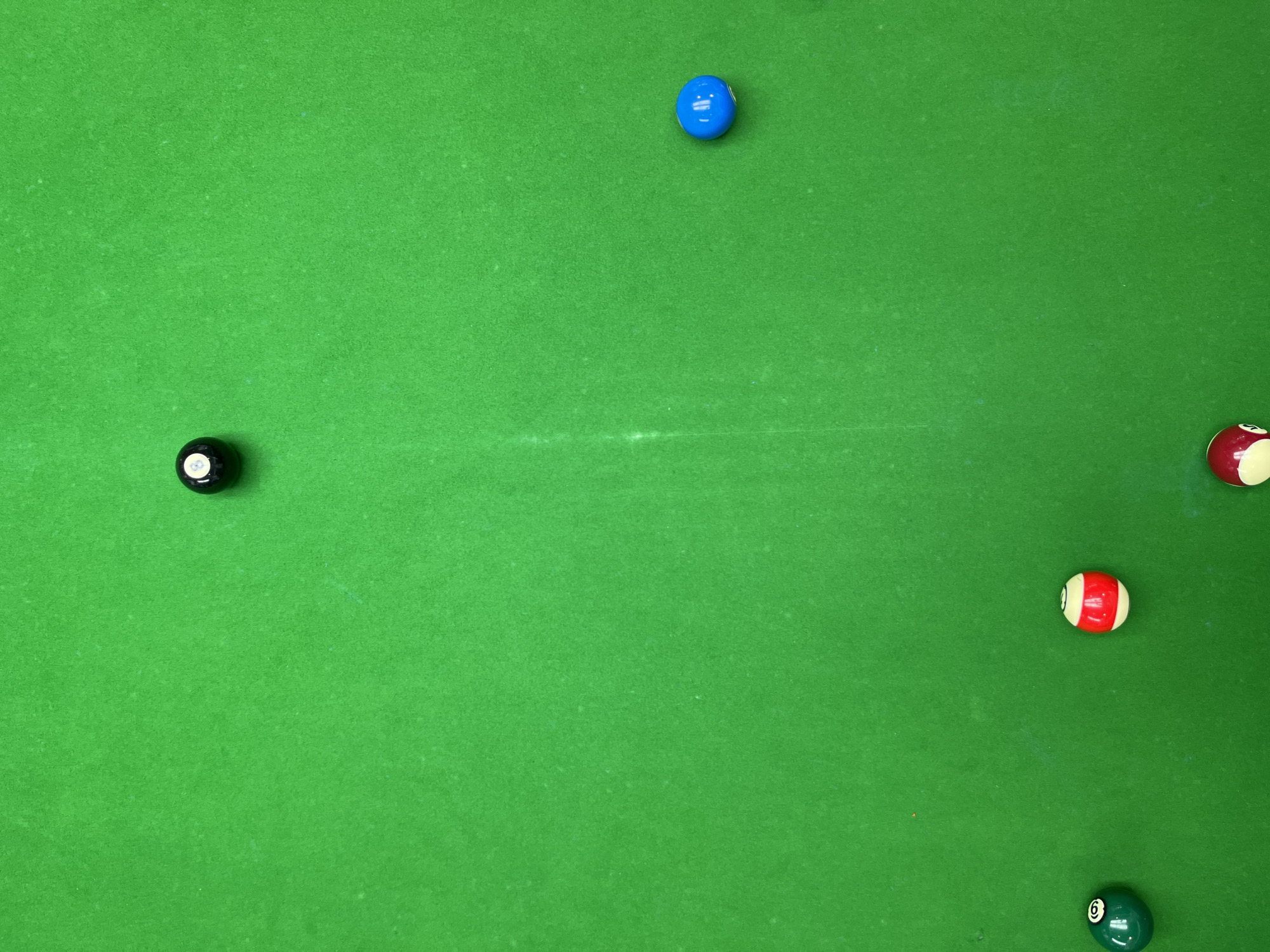 An overhead, full shot captures a vibrant green billiard table with six billiard balls scattered across its surface. The balls are of various colors and numbers, suggesting a game is in progress or has recently concluded.
The balls include a solid black 8-ball positioned on the left side of the table, a bright blue ball towards the top center, and a red and white striped ball below it. Further to the right, a red and white striped ball with the number 3 is visible, and another red and white striped ball with the number 7 is near the bottom right corner. A solid green ball with the number 6 is also present in the lower right quadrant.
The lighting is bright, indicating it is daytime, and the scene is indoors, typical of a billiard hall or a game room. The green felt of the table is smooth and shows some minor wear, common for a well-used billiard surface. There are no people visible in the frame, focusing all attention on the arrangement of the balls. The image is devoid of any discernible text other than the numbers on the billiard balls themselves. The setting is a generic billiard table, with no specific identifying features of the Daxing District, China location beyond the provided context.
