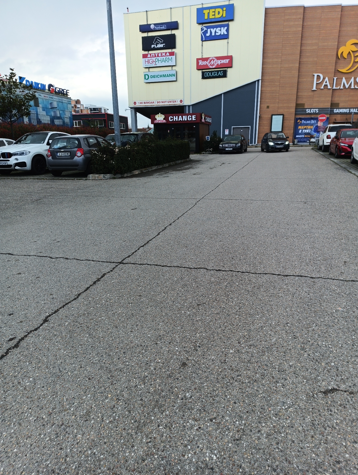 The image depicts a parking lot in front of a multi-tenant building, likely a shopping center, in Burgas, Bulgaria. Signage indicates the presence of businesses such as Tedi, Jysk, Техномаркет, Douglas, Postbank, Flair, Аптека High Pharm, and Deichmann. Several cars are parked in the lot, including a white BMW, a grey car, a dark sedan, and a black car. A sign for "Palms" is visible on a section of the building, indicating it houses gaming halls. The pavement is asphalt and shows signs of wear and cracking. A row of shrubs lines a portion of the parking lot near the storefronts. The sky appears overcast. A sign that says "CHANGE" can be seen on one of the storefronts.