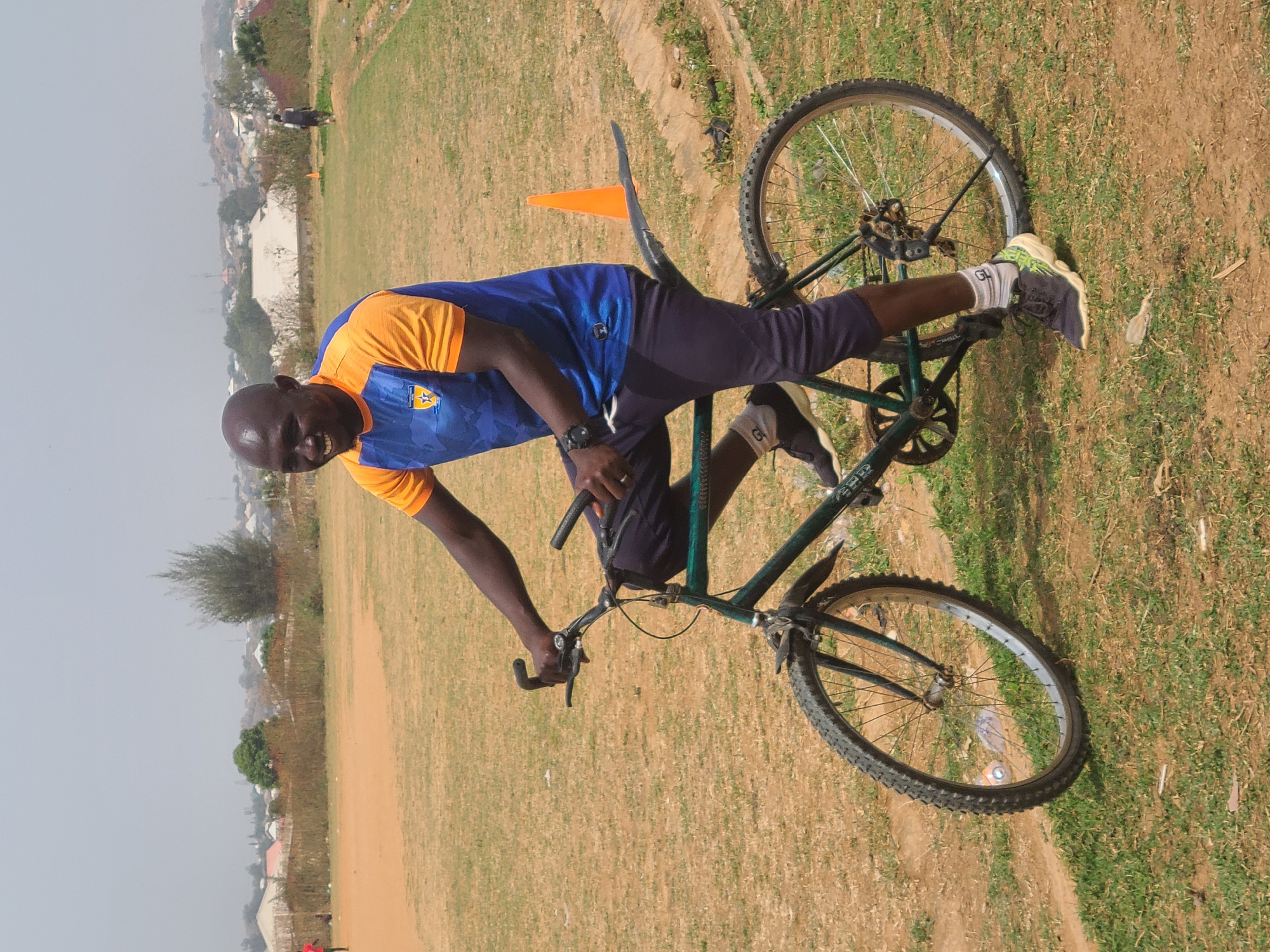 An adult male is positioned sideways on a dark green bicycle in an open grassy field. He is wearing a blue and orange sports jersey, dark leggings, and running shoes. His right foot is on the bicycle pedal, and his left foot is on the seat. The man is smiling and looking at the camera. In the background, there is an orange cone, and blurred structures suggest a built-up area. The location is Barikin Jirga Summa, Nigeria. The ground cover is mixed with green grass and brown soil, and there is scattered debris. The sky is overcast.