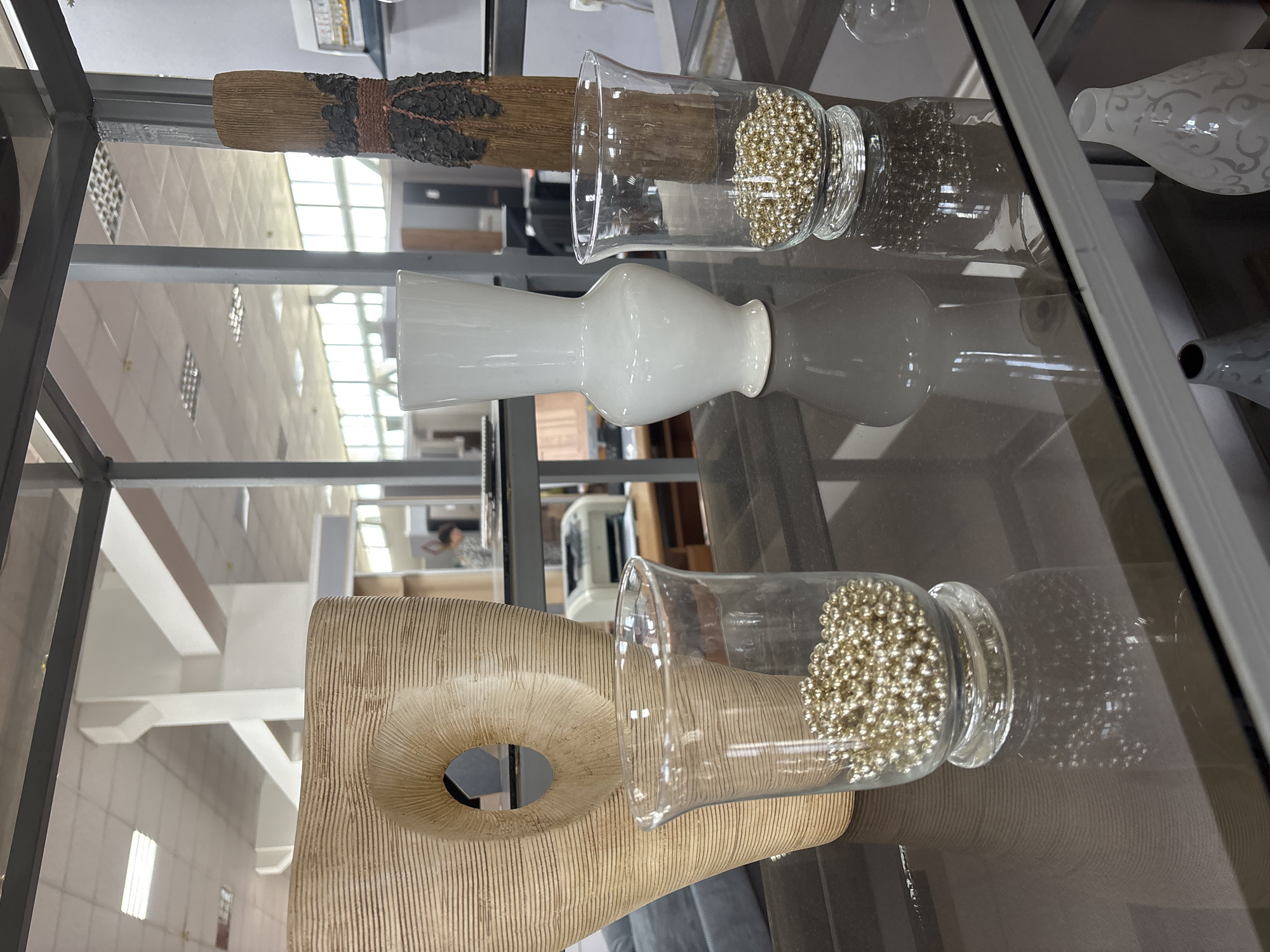 In an indoor retail environment, possibly a home decor store, three vases are displayed on a dark, reflective surface. The largest vase, on the left, is made of a textured, light-colored material and has an abstract, organic shape with a large oval opening. To its right, a tall, slender white vase with a narrow neck and wider base sits centrally. On the far right, a decorative object resembling a piece of wood wrapped in dark material and twine stands upright. In front of these vases, two clear glass vessels are placed. The left glass vessel contains numerous small, spherical metallic beads. The right glass vessel also holds a similar collection of metallic beads. The background reveals shelves and office-like furniture, including a printer, suggesting a commercial or office setting. The lighting is overhead and fluorescent.