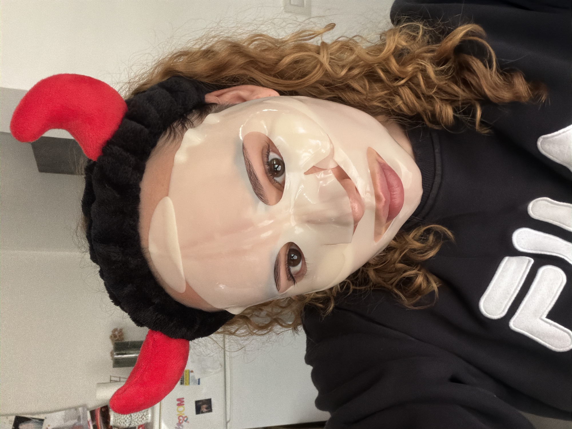 A person is indoors wearing a black headband with red devil horns and a sheet face mask. They are wearing a black hoodie with a white FILA logo. The environment appears to be a domestic interior, with a blurred refrigerator visible in the background. There is no discernible indication of the city Ramat Gan, Israel.