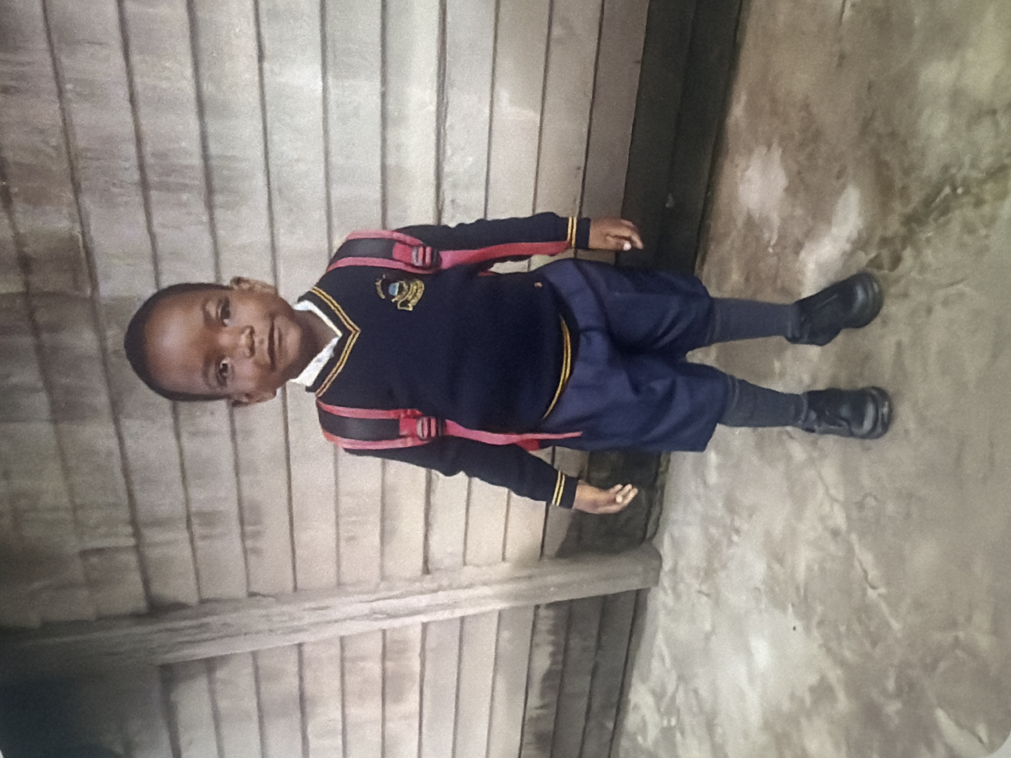 A young boy, dressed in a school uniform, stands in front of a wooden wall. The uniform consists of a navy blue sweater with yellow trim around the V-neck and cuffs, navy blue shorts, gray socks, and black shoes. He is wearing a red backpack with black straps. The background is a light-colored wooden wall with horizontal planks. The ground in front of him is a light gray, mottled concrete surface. The scene appears to be outdoors, possibly in a schoolyard or a similar environment.