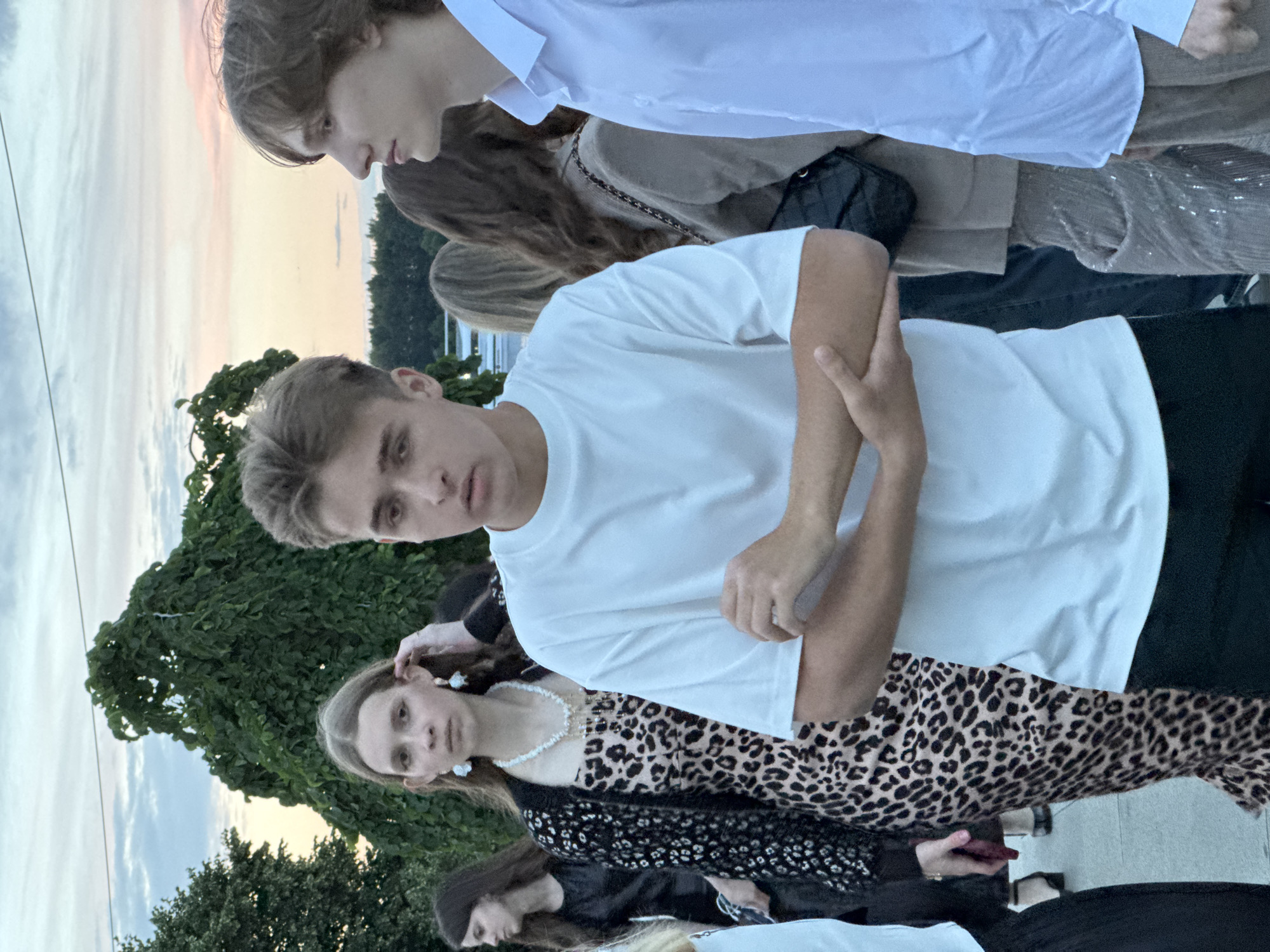 In an outdoor setting with a dusky sky, a group of young people are gathered. In the foreground, a young male with short, light brown hair, wearing a white t-shirt, stands with his arms crossed. To his right, another young male, also with light brown hair, is seen in profile, wearing a light blue collared shirt. To the left of the first male, a young female with long brown hair is visible, wearing a leopard-print dress and a black patterned cardigan, adorned with a pearl necklace and large white earrings. Further to the left, another young female with dark hair is partially visible. In the background, a large, dense green tree dominates the scene, with a blurry body of water and trees in the far distance. The ground appears to be paved.