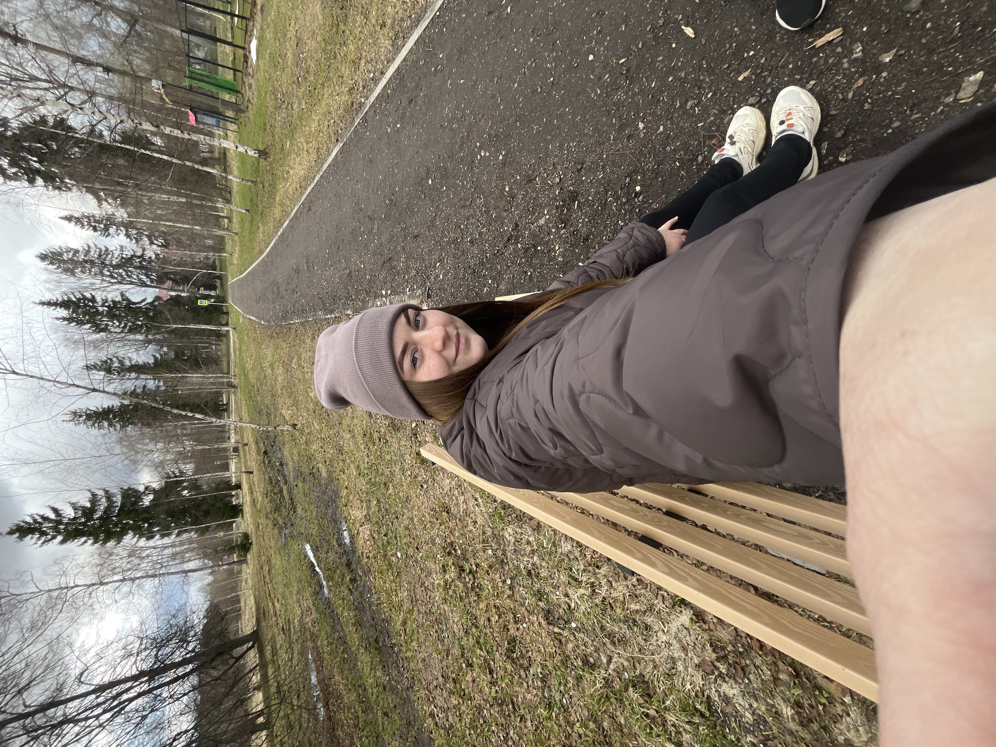 A young woman sits on a wooden bench along a paved path in a park, likely in Vysokovsk, Russia. The woman is wearing a gray beanie, a brown puffer jacket, and black pants. She has white sneakers on her feet. She is looking at the camera and smiling. The background of the image includes bare trees, grass, and another path. The ground is covered in grass and some dried leaves. The bench is made of light-colored wood.