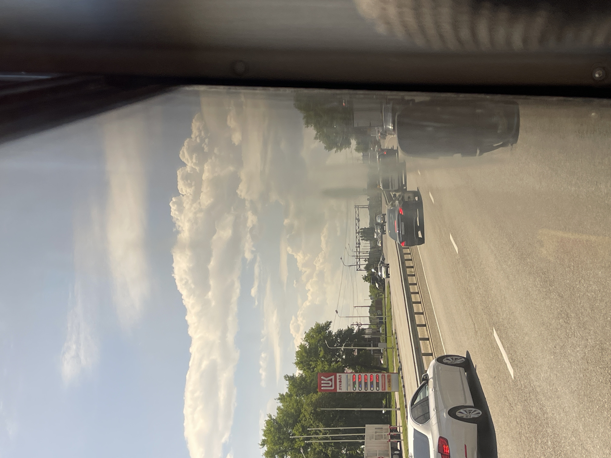 The image depicts a view from inside a vehicle, likely on a highway, with the camera oriented vertically. The scene is a roadside with a clear sky filled with large, white cumulus clouds, suggesting a sunny day. The road is asphalt with visible white lane markings, and it appears to be a multi-lane highway with several cars driving in the same direction. Trees line the side of the road, and in the distance, there are more cars and what appear to be streetlights and possibly buildings.
A prominent object in the scene is a gas station sign for "ЛУКОЙЛ" (LUKOIL), a Russian multinational energy corporation. The sign displays fuel prices, with various types of fuel listed. Next to the LUKOIL sign, there are more structures associated with a gas station, including pumps.
The activity taking place is vehicular traffic on a highway. The presence of the gas station indicates it's a location where drivers might stop for fuel. The time of day appears to be daytime, given the bright sunlight and clear sky, likely late morning or afternoon. The overall impression is a typical scene of travel on a highway in a suburban or semi-rural area of Russia. There are no people clearly visible in the image. The mood is one of everyday travel.