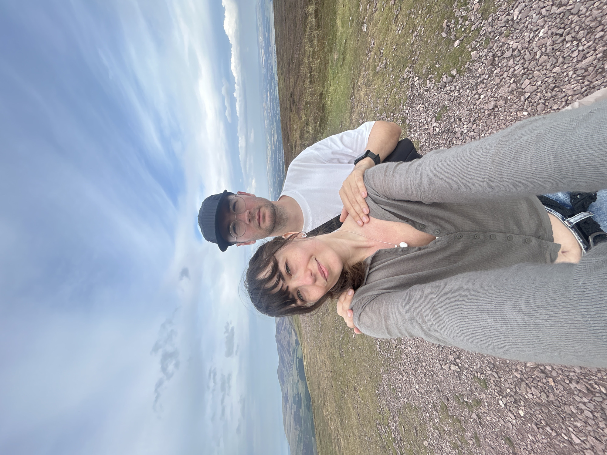 A low-angle, selfie-style photograph captures a couple standing on a rocky, grassy hillside with a panoramic view of undulating, green hills and a distant town under a partly cloudy sky. A woman is in the foreground, smiling and looking at the camera, with a man behind her with his arm around her shoulder. The woman is wearing a gray, button-up cardigan and denim shorts, while the man is wearing a white t-shirt, glasses, and a black baseball cap. The setting appears to be a rural or mountainous area, possibly within or near the city of Penicuik, United Kingdom, given the resemblance to the Scottish landscape. The lighting is soft, suggesting an overcast day.