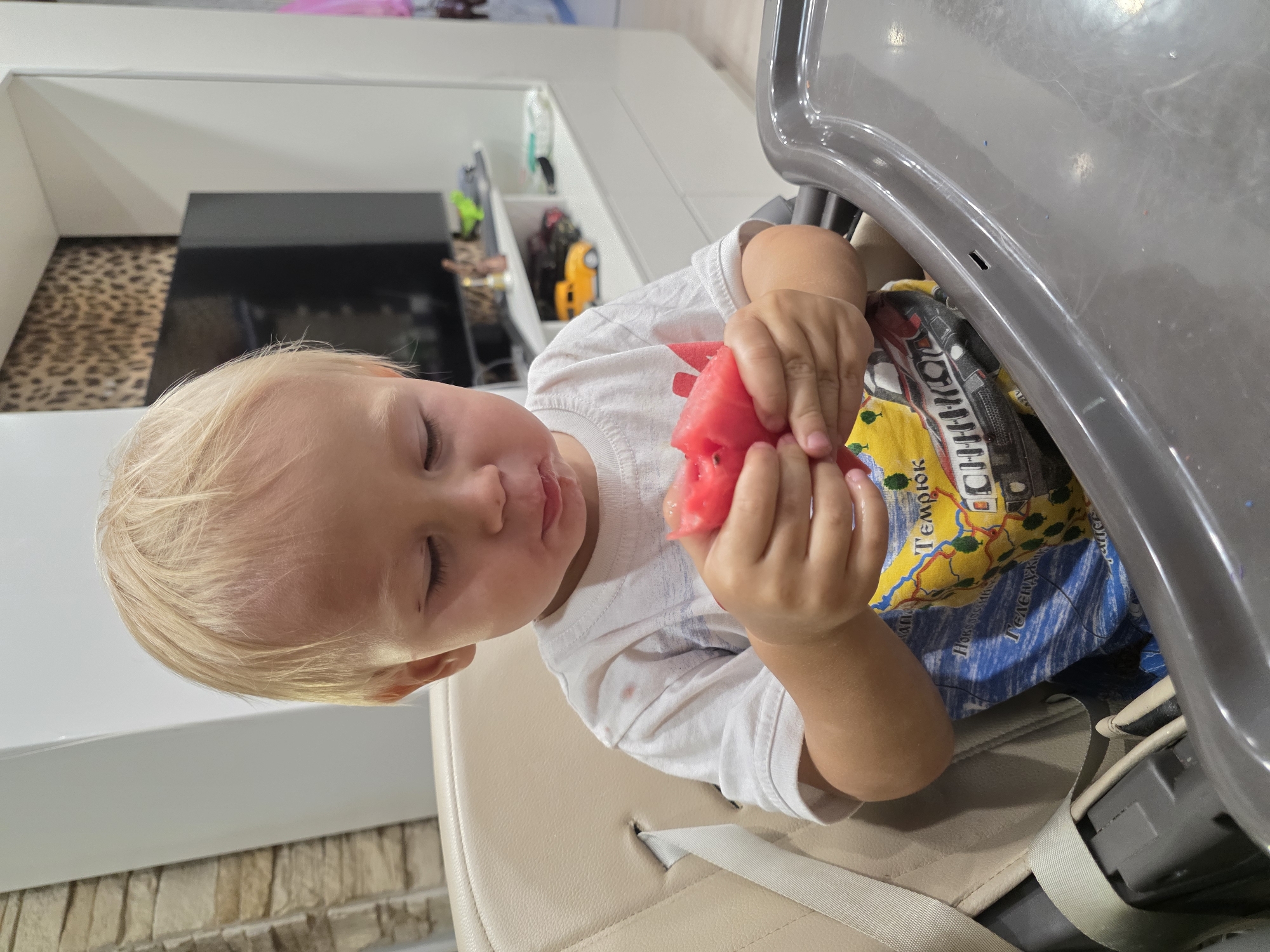 A young boy with blonde hair is seated in a high chair, holding and eating a slice of watermelon. He is wearing a white t-shirt with a colorful graphic print. The high chair is beige with a grey tray. In the background, there is a television set with a leopard-print wallpaper above it. A shelf unit with various items, including a yellow toy car, is visible. The overall setting appears to be a domestic environment, possibly a dining area within a home. There is no specific indication of a location within Saint Petersburg, Russia.