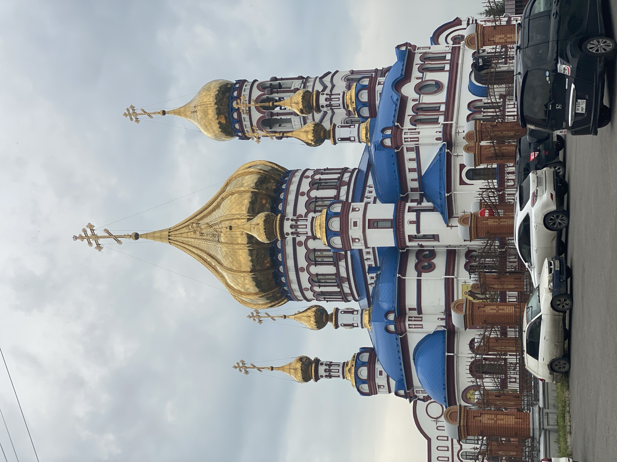 An imposing orthodox church with multiple gold-domed towers, topped with golden crosses, and bright blue roofs stands against a cloudy sky. The white church features red trim and arched windows. In the foreground, several cars are parked on a paved area in front of a brick fence surrounding the church. The location is identified as Novokuznetsk, Russia.