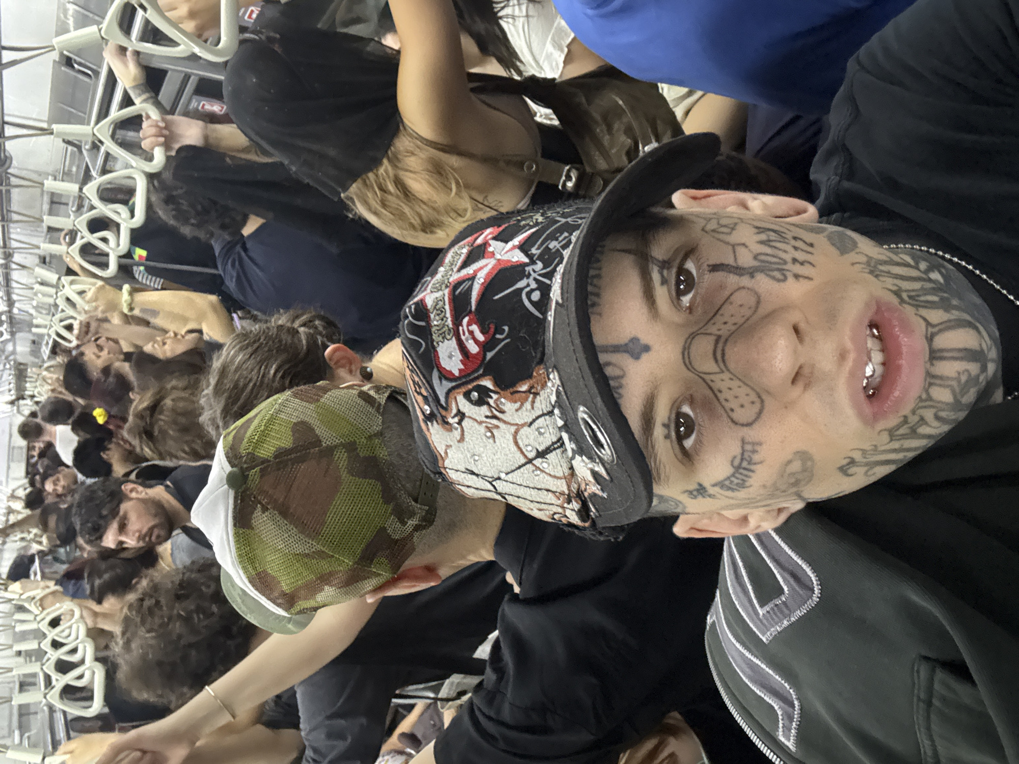 A crowded subway car in Buenos Aires, Argentina. People are standing and holding onto overhead grab bars. A man with multiple face tattoos and silver dental grills is looking at the camera. He is wearing a black baseball cap with designs on it. Another man beside him is wearing a camouflage cap.