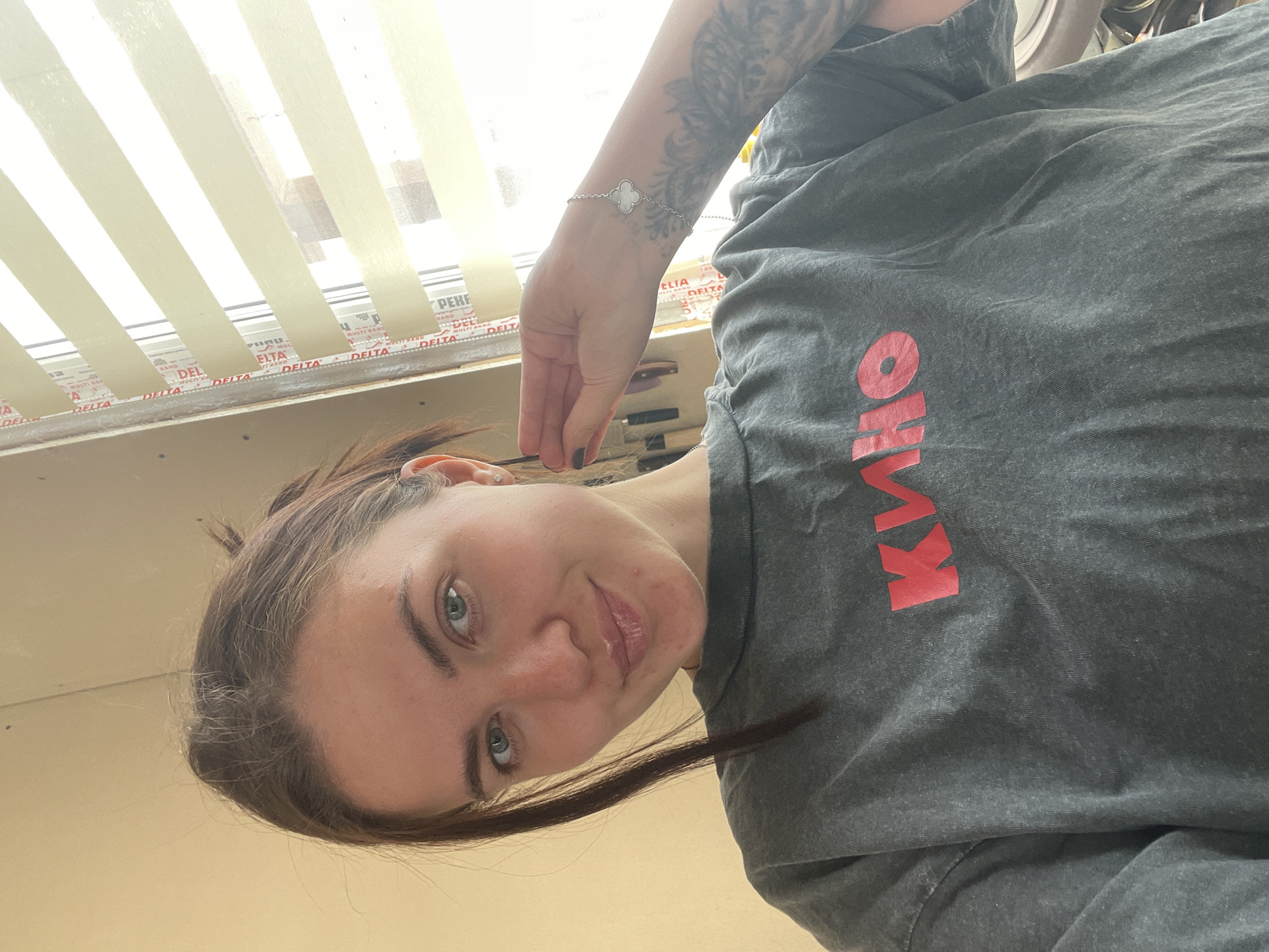 Here is a matter-of-fact description of the image:
A woman with fair skin, light brown hair, and light-colored eyes is looking down at the camera. She is wearing a gray t-shirt with the word "КИНО" printed on it in red. On her left wrist, she is wearing a silver bracelet with a clover charm. Part of her arm is visible, showing a tattoo.
In the background, there is a window with white blinds. The window frame has the word "DELTA" printed on it repeatedly. Some construction-related items are visible near the window, but they are out of focus.
There is not enough information to determine the precise location in the city of Мосметаллоконструкция Онт, Russia.