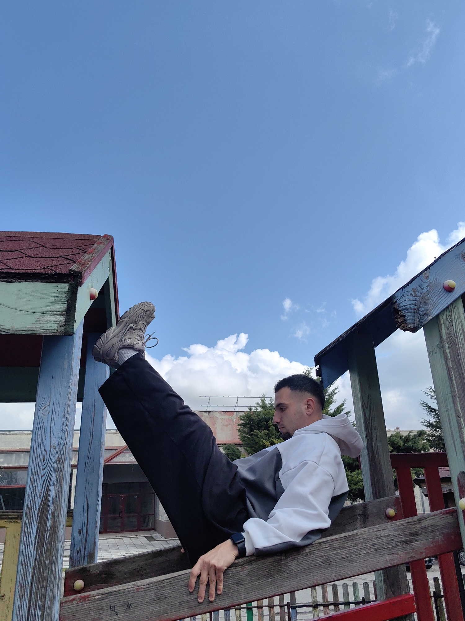 A low-angle, wide shot depicts a man in an unusual posture on a playground structure on a bright day with a blue sky and scattered white clouds. The man, positioned horizontally between parts of the structure, appears to be sitting on one wooden ladder-like section while his legs are extended vertically towards the top of the frame. He is wearing a grey and white hoodie, dark pants, and light-colored sneakers. A watch is visible on his left wrist. The weathered playground equipment is made of painted wood in shades of blue, red, and green. Buildings are visible in the background, including some with red roofs. The location is Shumen, Bulgaria.