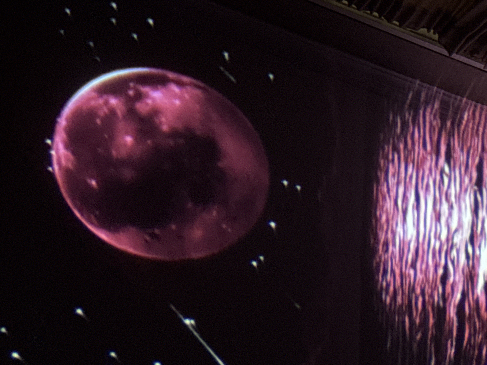 The image shows a projection of a cosmic scene onto a surface, likely a screen or a wall. A large, reddish-purple celestial body, resembling a planet or moon, dominates the upper portion of the frame. It is illuminated and shows some surface texture. Scattered around it are numerous small, bright white or light blue star-like points, some appearing as streaks suggesting movement. Below this celestial display, there is a rippling surface of water reflecting the reddish-purple light of the planet, creating an abstract pattern of light and color on the water. The bottom right of the image shows a dark, textured edge, possibly a frame or part of the viewing environment. There are no people or identifiable landmarks of Tbilisi visible.