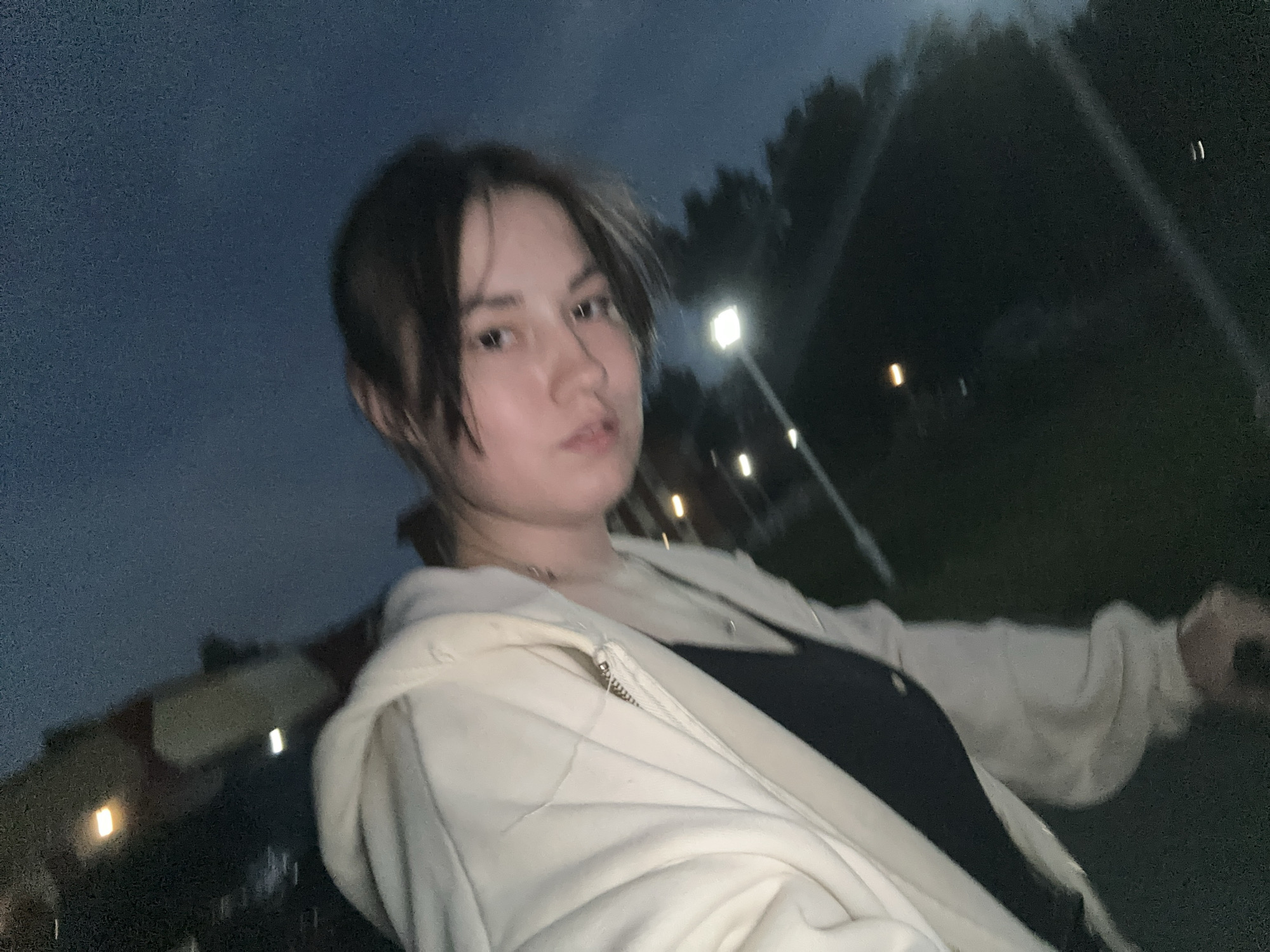 The image is a dimly lit, tilted selfie taken outdoors at dusk. A young person with dark hair and light skin is in the foreground, wearing a light-colored hoodie over a dark shirt. Their face is turned towards the camera, with their eyes looking slightly to the side. In the background, out-of-focus streetlights and trees are visible against a dark blue sky. The setting appears to be a park or public area, possibly near a road or pathway, illuminated by artificial light. There is no specific visual information to indicate the exact location within the city of Chaikovsky, Russia.