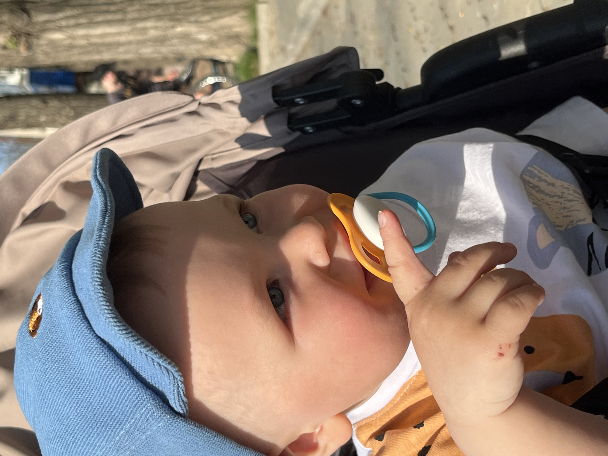 A baby with fair skin, wearing a blue cap and a shirt with an orange pattern, lies in a stroller with a beige canopy. The baby has a pacifier in their mouth and is holding the pacifier with their right hand. There is a black stroller handle visible. In the blurred background, there appears to be an outdoor urban scene. The location of the photo is not possible to tell using visual features only.