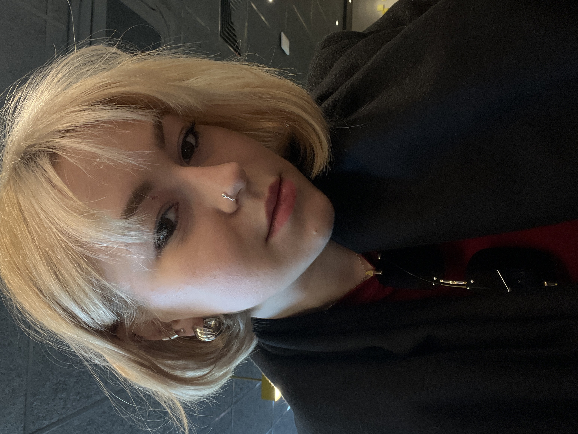 This close-up portrait, taken in Odesa, Ukraine, features a young individual with short, light blonde hair and light skin, looking directly at the camera with a neutral to slightly contemplative expression. The image is significantly tilted to the left. The subject has dark eyes, a small silver hoop nose piercing, and multiple silver-toned earrings, including an ear cuff on the visible ear. They are wearing a dark, possibly black, jacket or coat over a red garment. A pair of sunglasses with gold-colored frames is resting on the jacket on the right side of the frame. The background is dimly lit, featuring dark, textured surfaces with subtle geometric patterns and soft, diffused light sources, suggesting an indoor public setting like a cafe or restaurant. The overall lighting is low, creating a somewhat intimate atmosphere. No visible text is present in the image.
