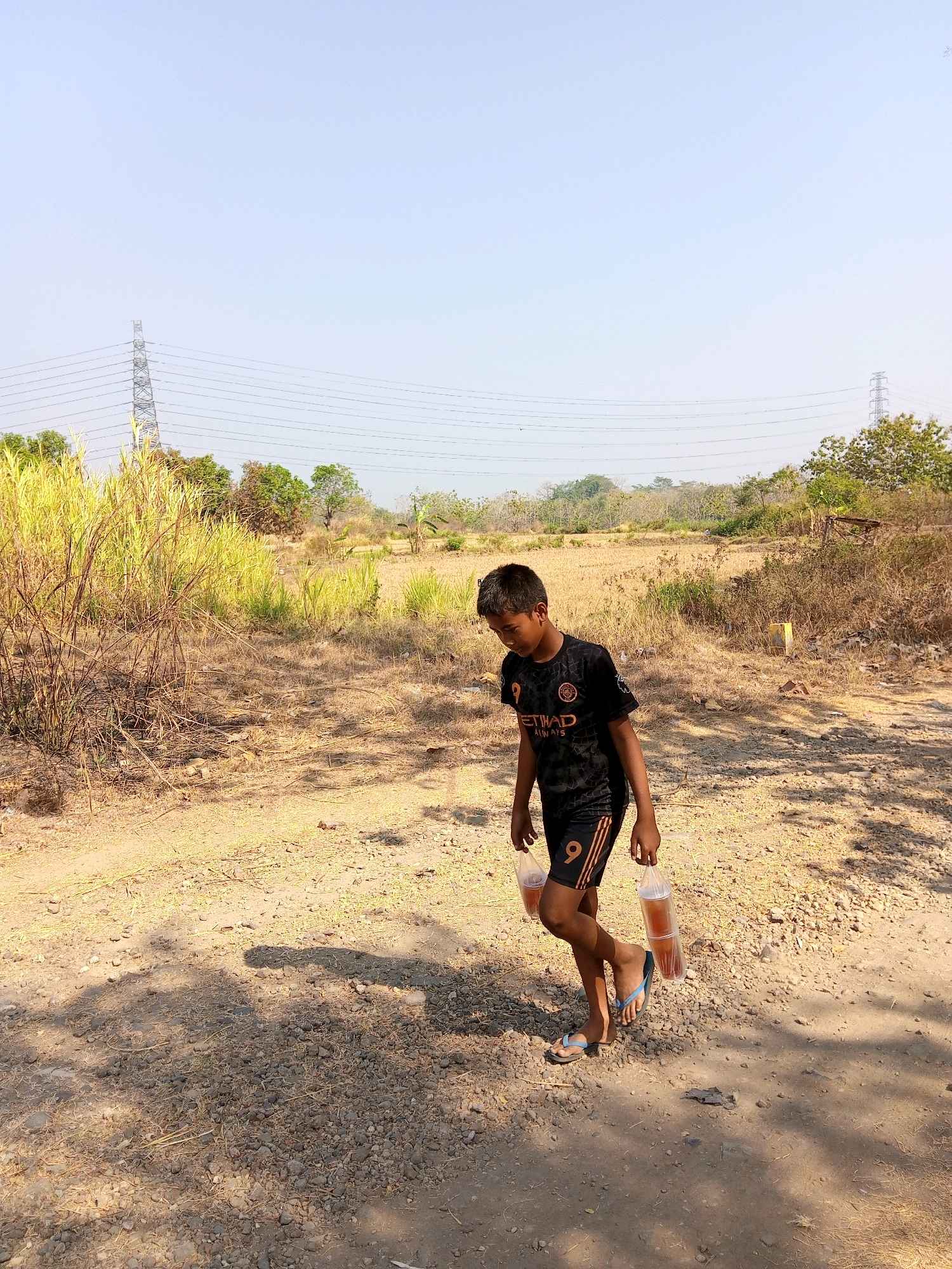 A young boy walks on a dirt path in Sendangijo, Indonesia, carrying two plastic bottles filled with a dark liquid. He is wearing a black t-shirt with orange and white lettering and a number 9 on the sleeve, and black shorts with orange stripes. He is also wearing blue flip-flops. The path is dry and dusty, with sparse vegetation and some scattered rocks. In the background, there is a field of dry grass, trees, and a power line tower. The sky is clear and blue.